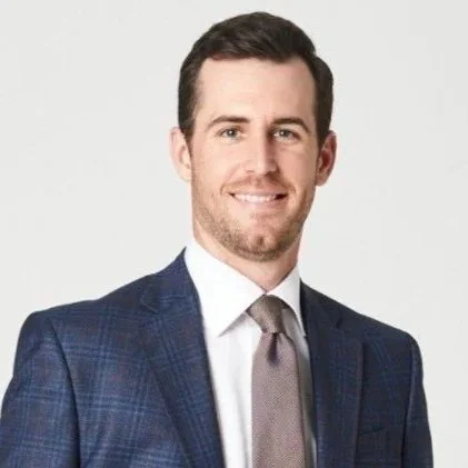 A smiling man with light brown hair and a beard, dressed in a navy suit, white shirt, and navy tie, in front of a neutral background.