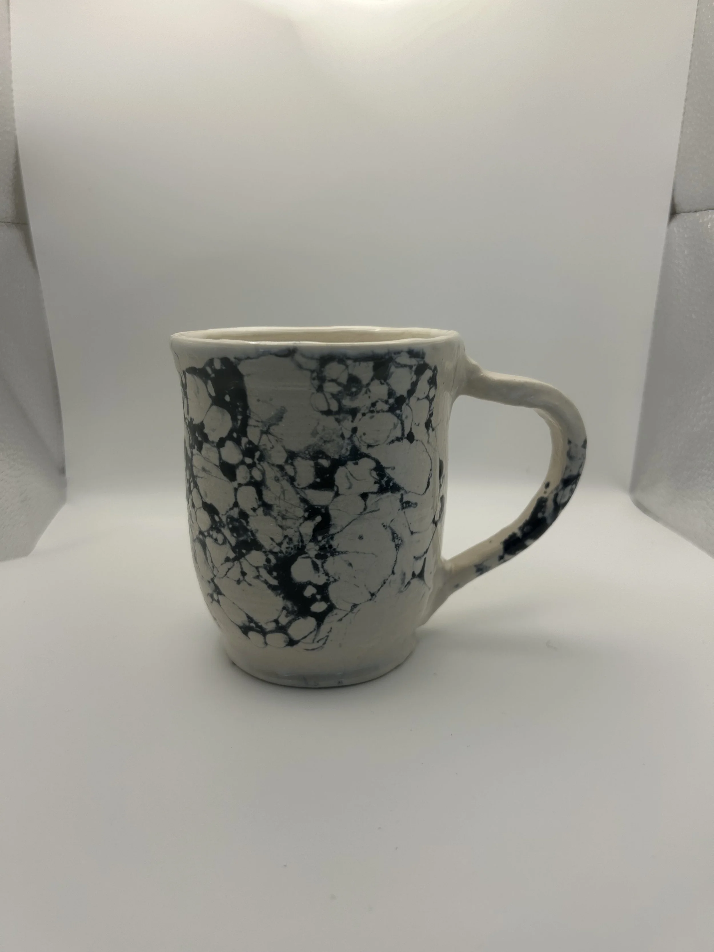 A ceramic mug with a white base and black marble-like veining pattern, placed on a white background with gray foam padding on the sides.