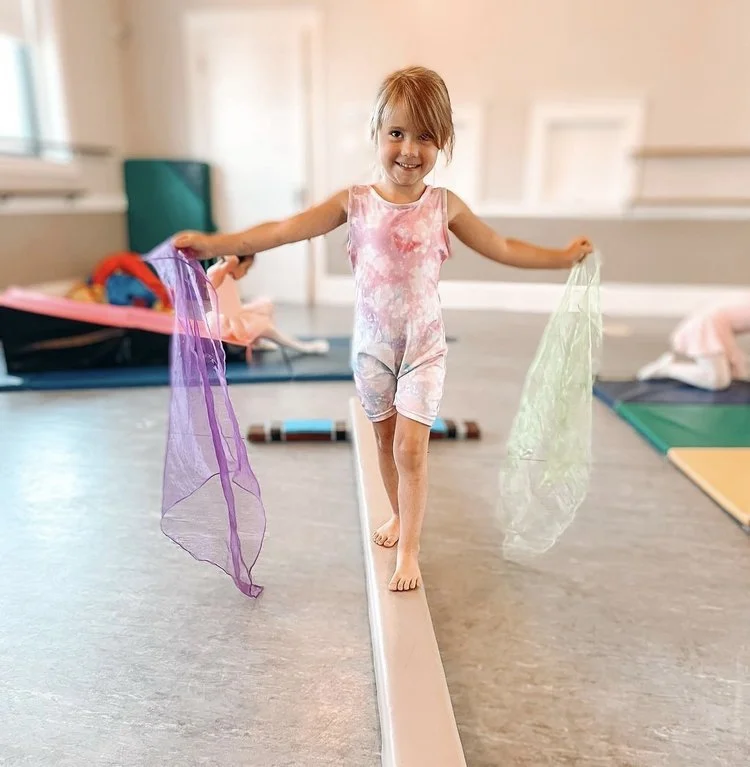 A young girl in a pink and white tie-dye dress balancing on a narrow balance beam in a playroom, holding colorful scarves, with mats and toys in the background.