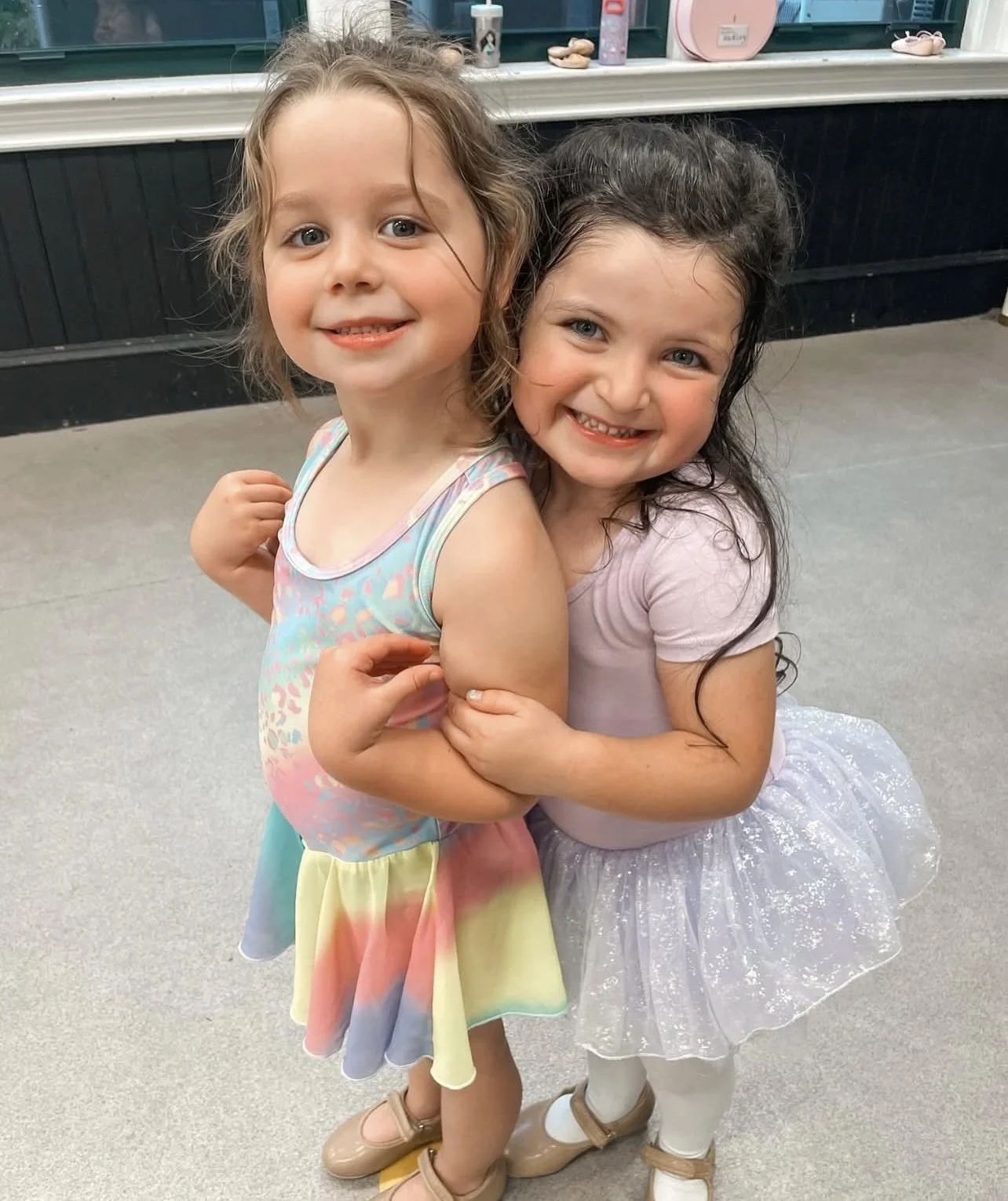 Two young girls smiling and hugging each other indoors, one wearing a pastel rainbow-colored dress, the other in a pink top and sparkly skirt.
