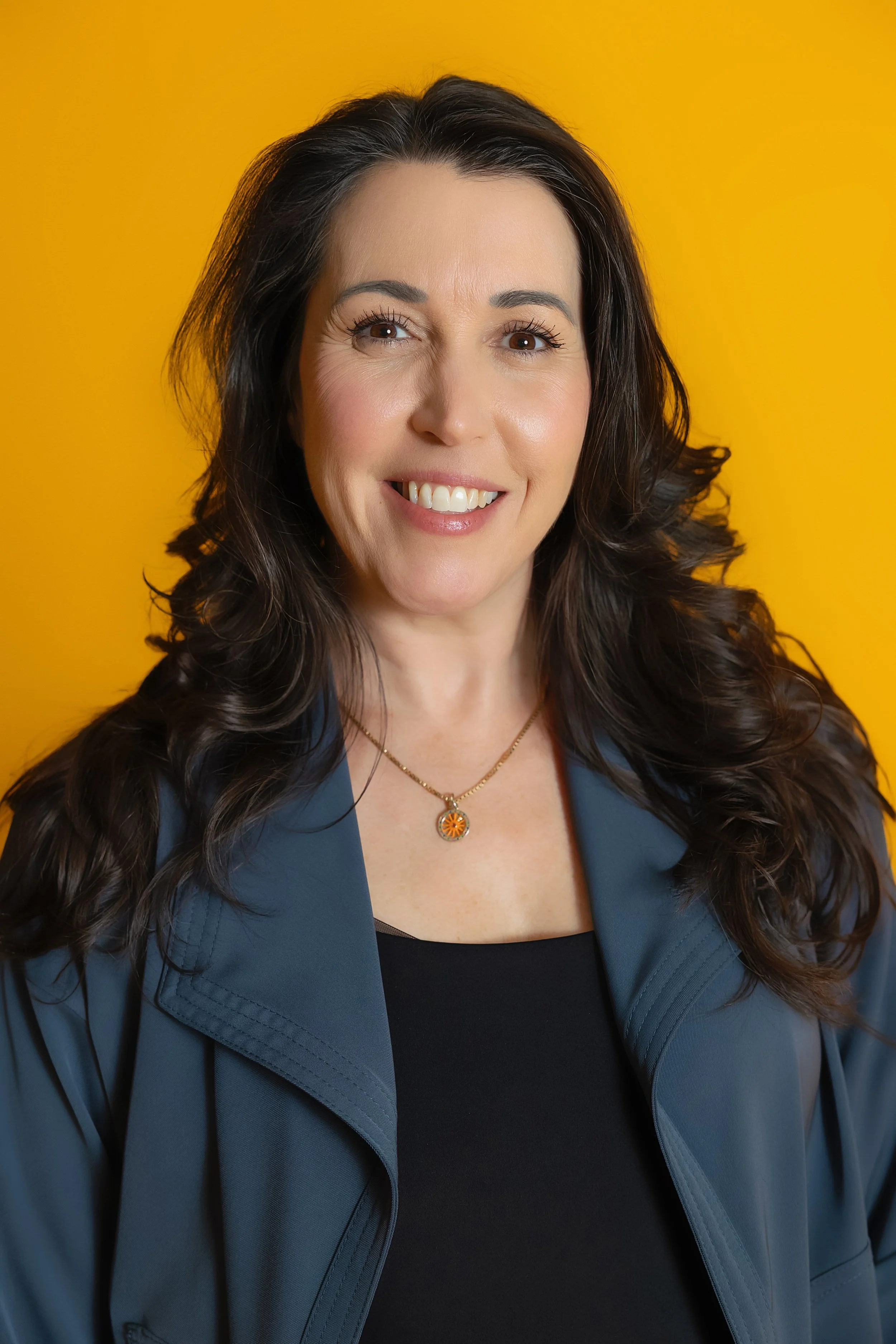 Portrait of a smiling woman with long dark hair, wearing a black top, blue jacket, and gold necklace, against a yellow background.