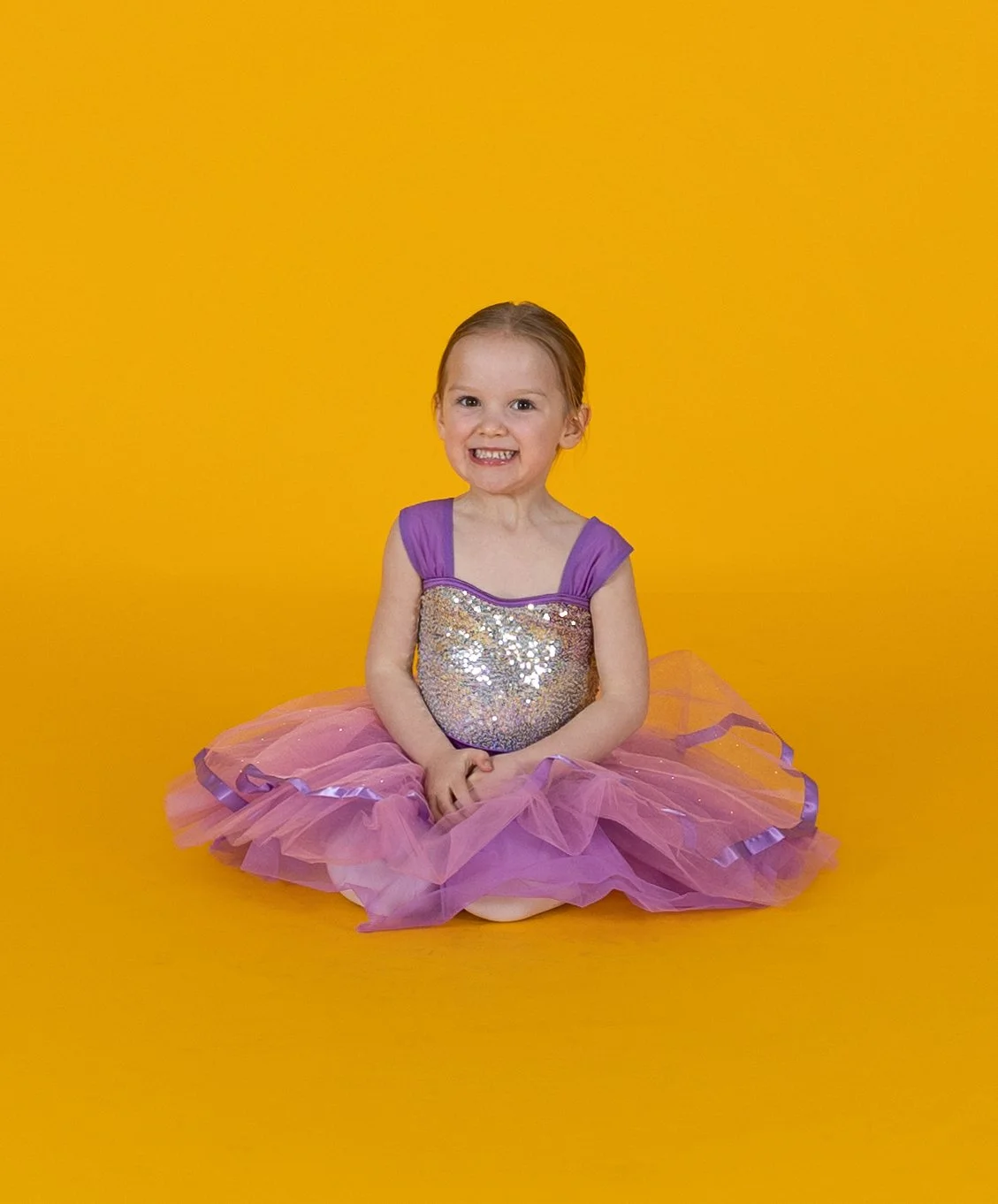 A young girl with brown hair in a purple and pink glittery dress, sitting on the floor against a yellow background, smiling with teeth showing.