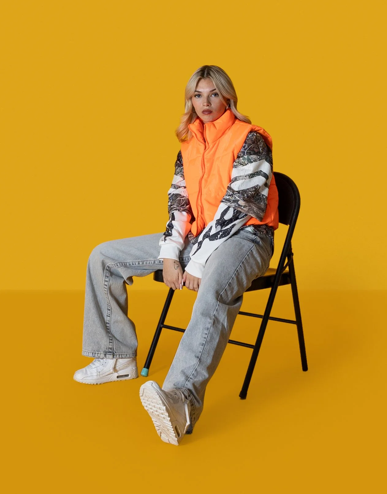 A woman sitting on a black chair against a yellow background, wearing a bright orange padded vest, a long-sleeved printed shirt, gray jeans, and white sneakers.