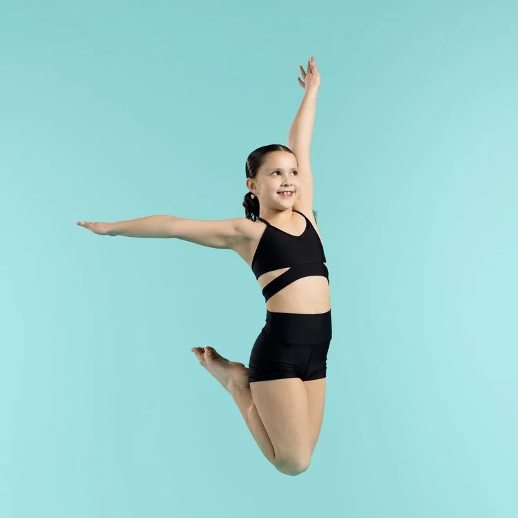 A young girl in black athletic wear jumping with arms outstretched and a smile against a light blue background.
