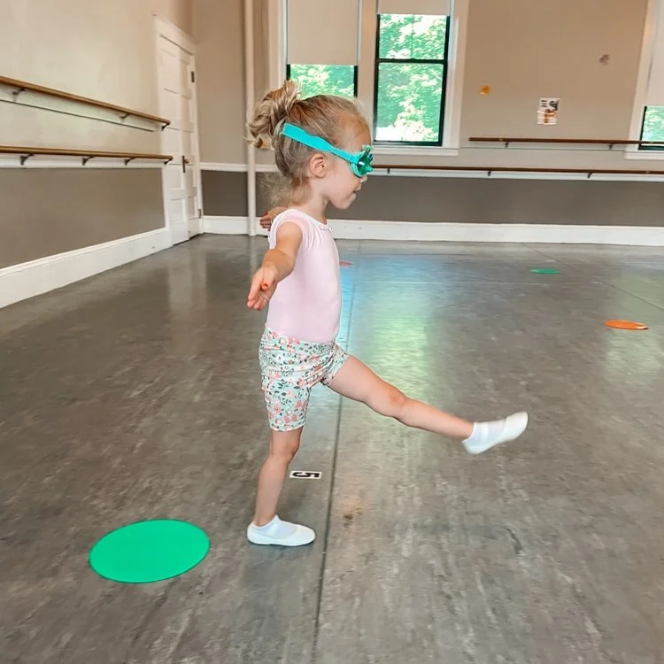 Young girl in a dance studio wearing swimming goggles, balancing on one leg with arms outstretched, surrounded by colored circular markers on the floor.