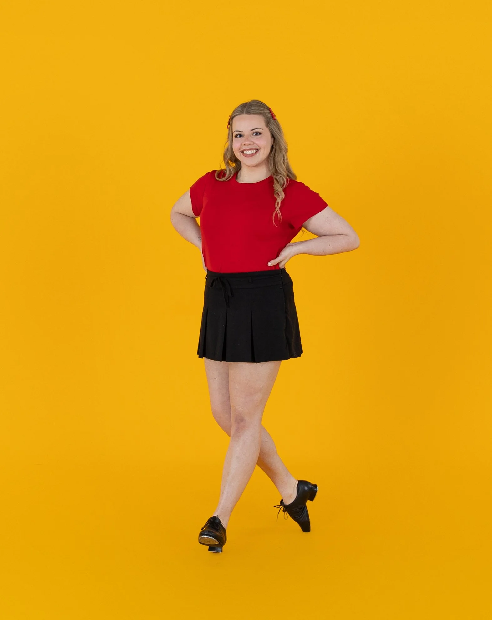 Young woman with blonde hair, wearing a red t-shirt and a black plaid skirt, standing with one foot slightly crossed over the other and smiling against a bright yellow background.
