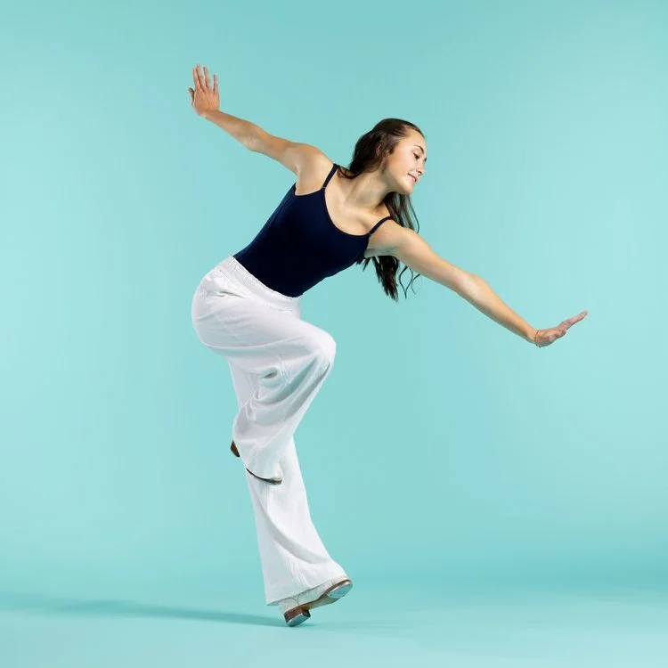 A woman wearing a navy tank top and white wide-leg pants on roller skates poses with arms extended on a light blue background.