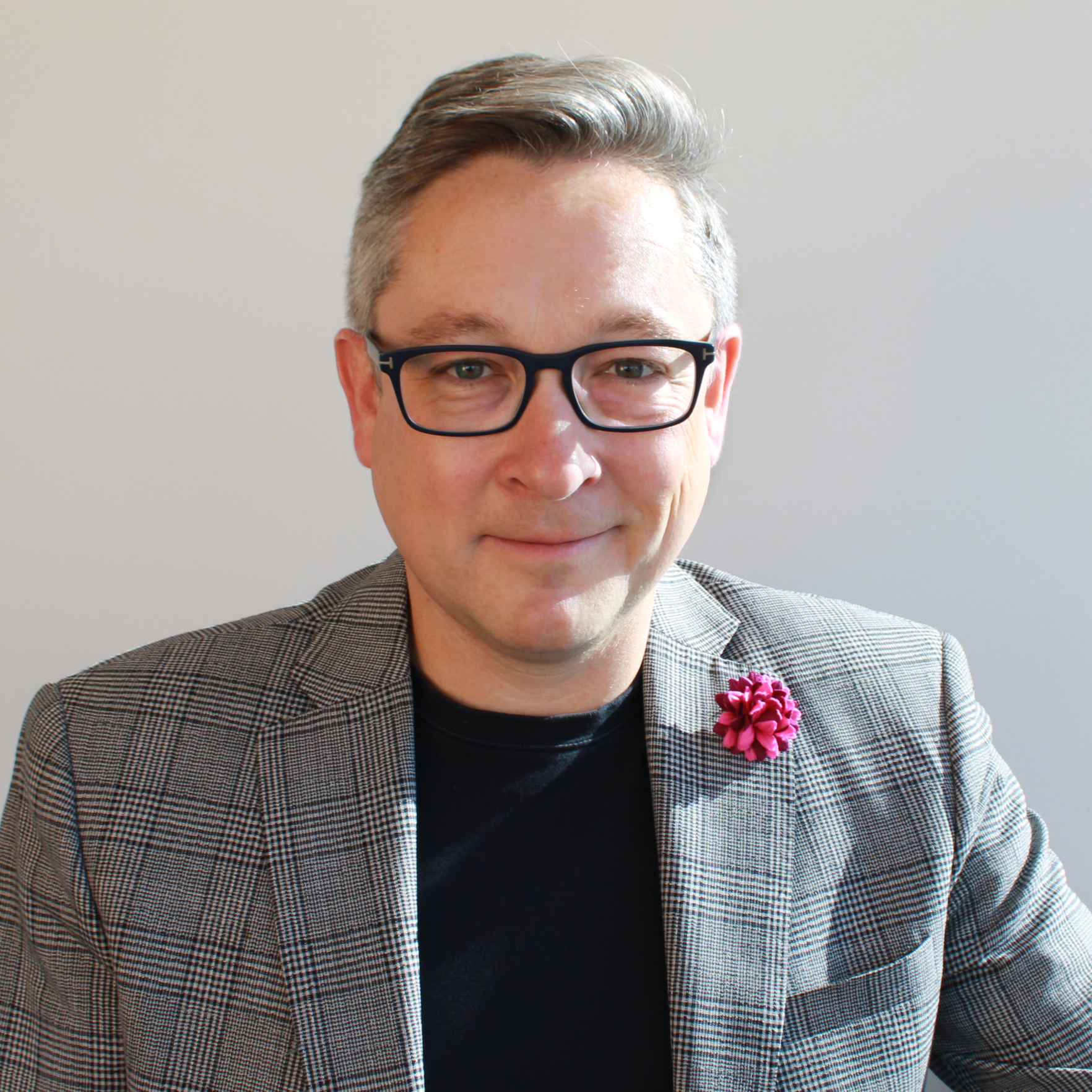 A man with short gray hair, glasses, and a checked blazer wearing a pink flower on his lapel, smiling at the camera against a plain background.