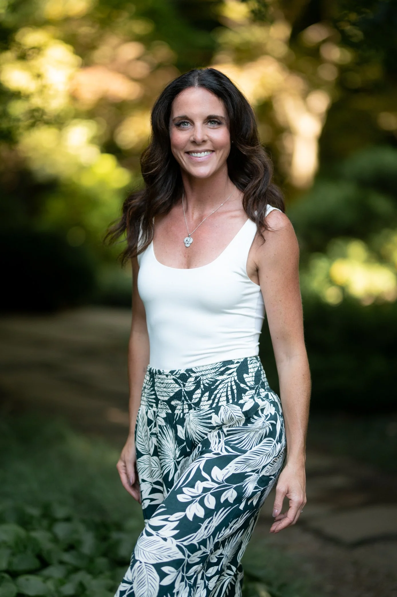 A woman with dark brown, wavy hair smiling outdoors in a natural setting, wearing a white tank top and black-and-white floral patterned pants.