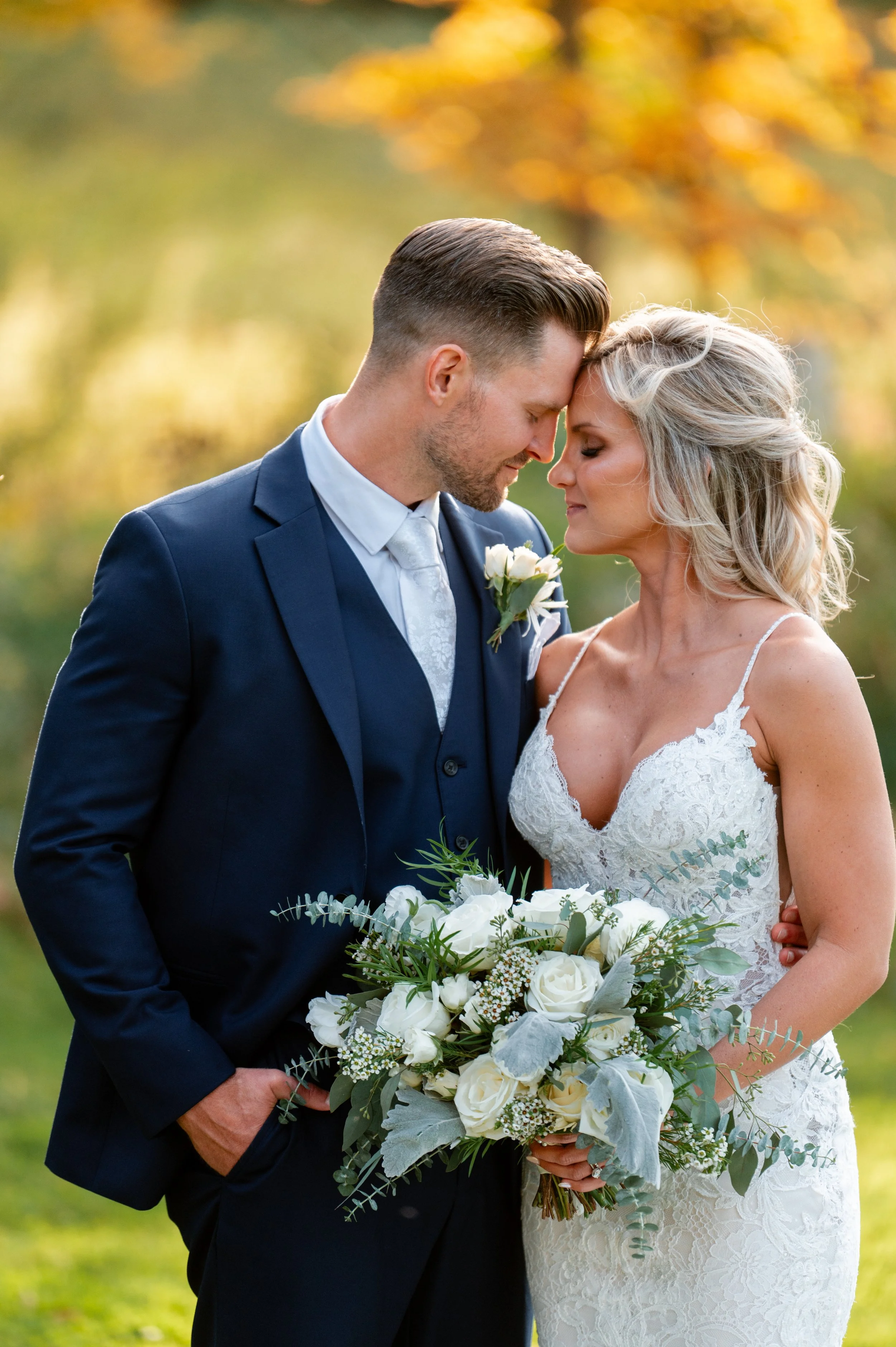 A bride and groom stand close with foreheads touching, in a park with fall foliage, the bride holding a bouquet of white roses and greenery.