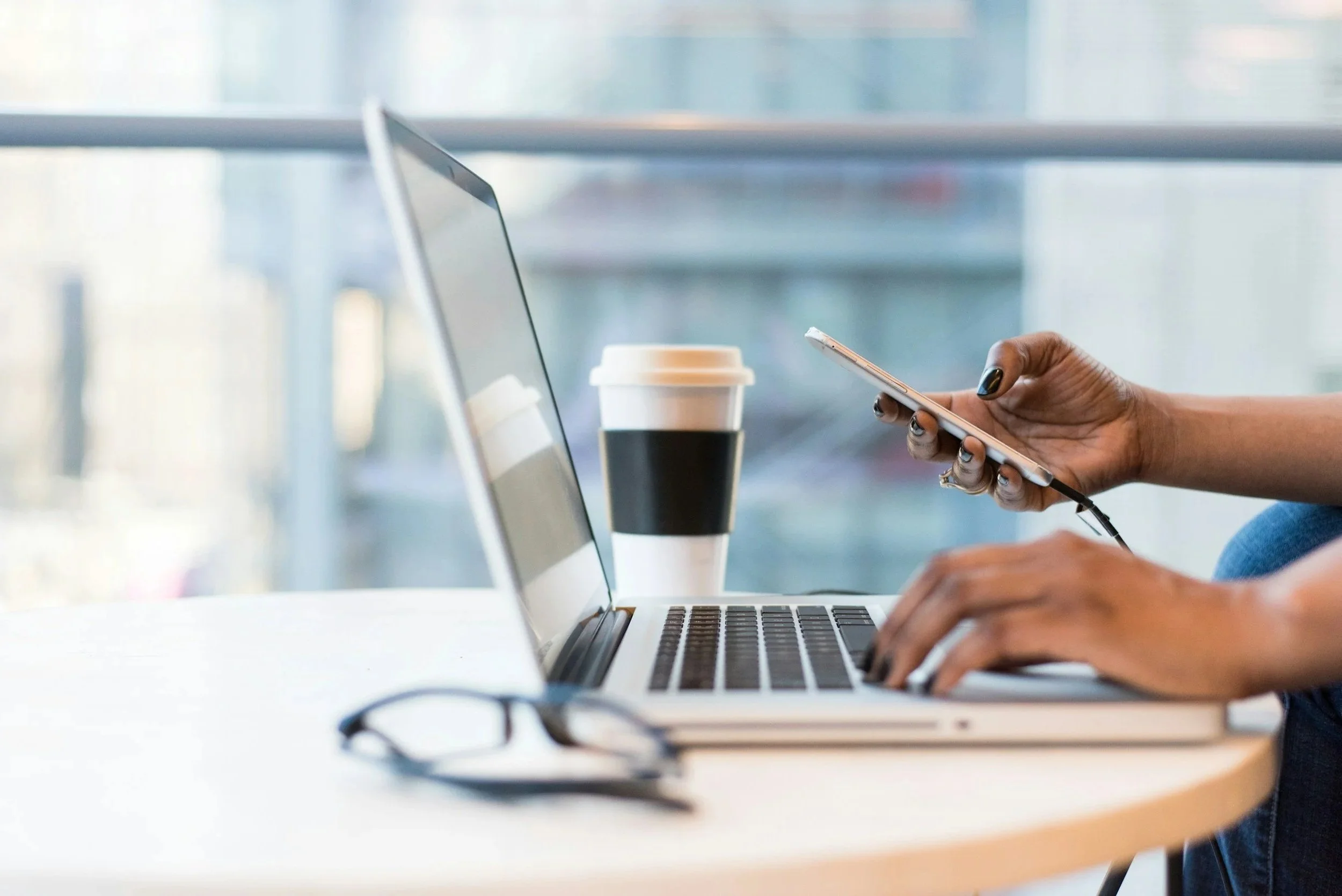 Une personne utilise un téléphone portable à côté d'un ordinateur portable sur une table, avec une tasse de café et des lunettes en premier plan.