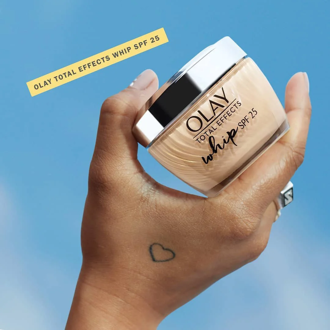 A hand holding a jar of Olay Total Effects Whip SPF 25 moisturizer against a blue background. The jar has a silver lid and the label shows the product name. The hand has a small heart tattoo on the wrist and is wearing a ring on the finger.