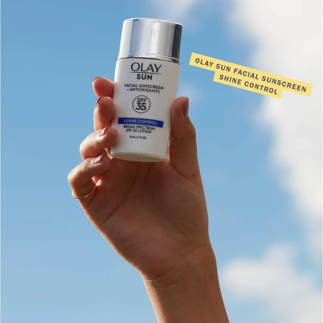 Hand holding a bottle of Olay Sun Facial Sunscreen Shine Control SPF 35 lotion against a blue sky with some clouds.