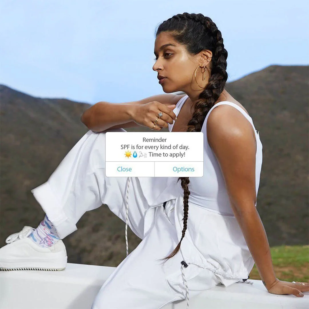 A woman with long braided hair sitting outdoors on a white surface in a mountainous area, wearing a white sleeveless jumpsuit, gold hoop earrings, and white sneakers with pastel-colored socks, while a notification about SPF sunscreen appears in front