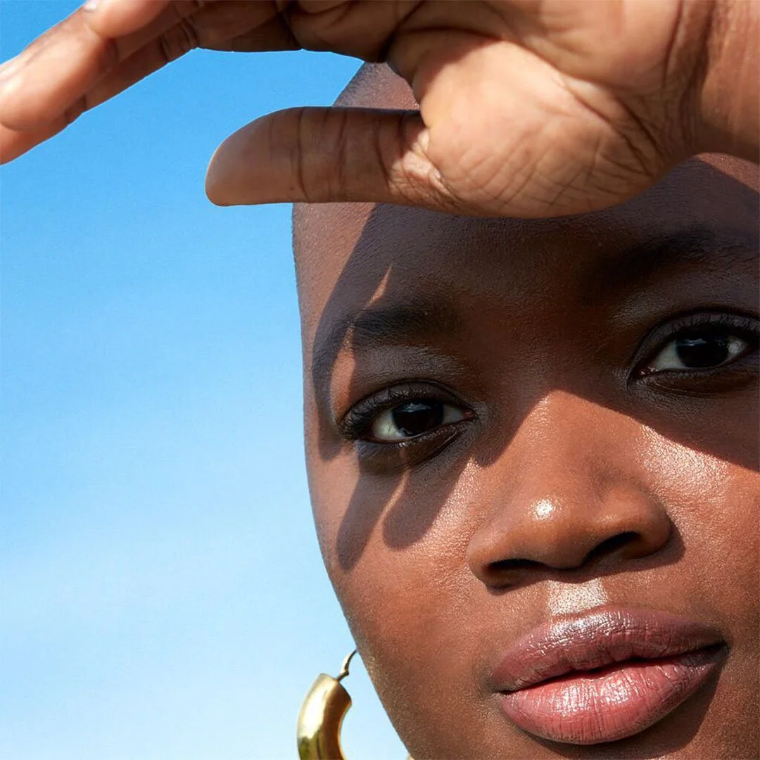 Close-up of a woman with dark skin, short hair, and a gold hoop earring, shielding her eyes from the sun with her hand against a clear blue sky.
