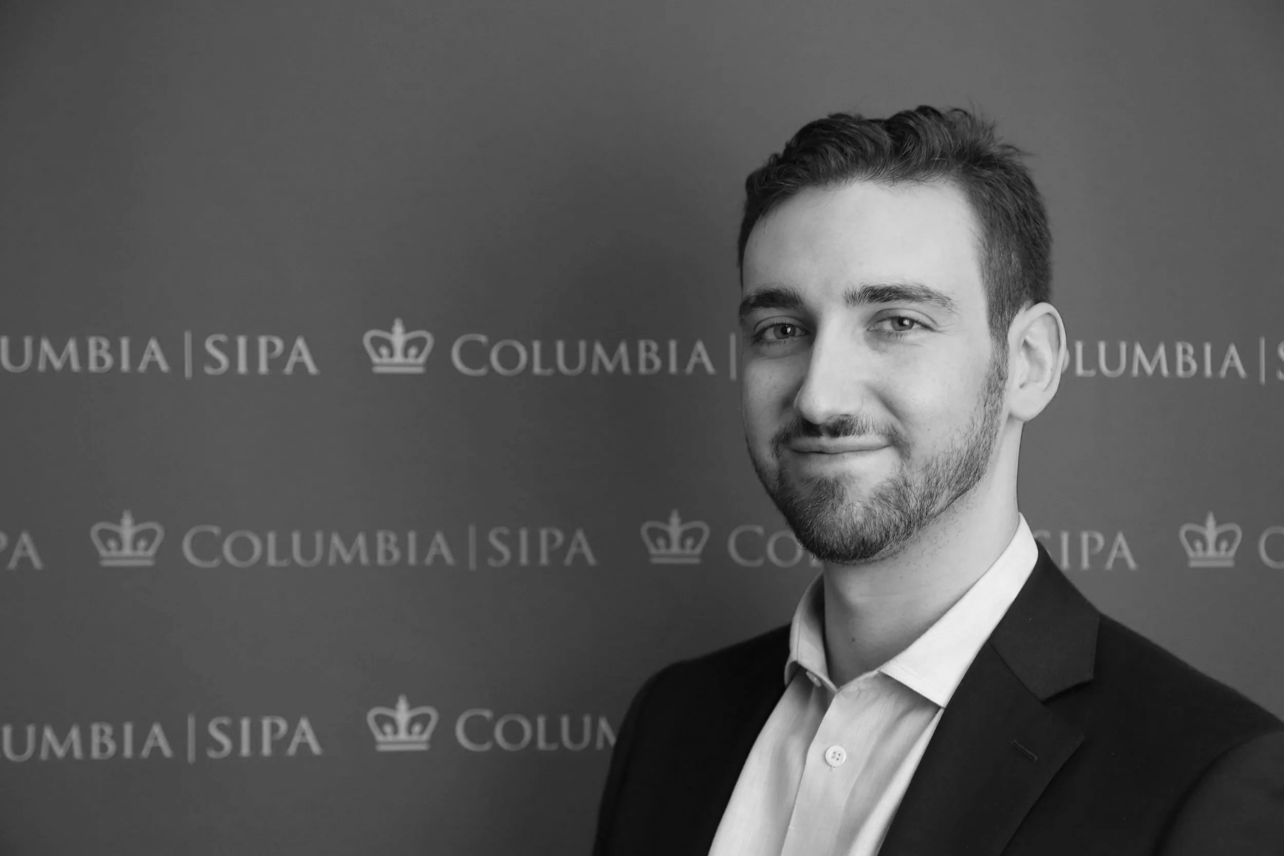 A young man with short brown hair, a beard, wearing a light blue dress shirt and a dark blazer, standing in front of a blue background with white text and logo that reads 'Columbia SIPA' and a crown symbol.