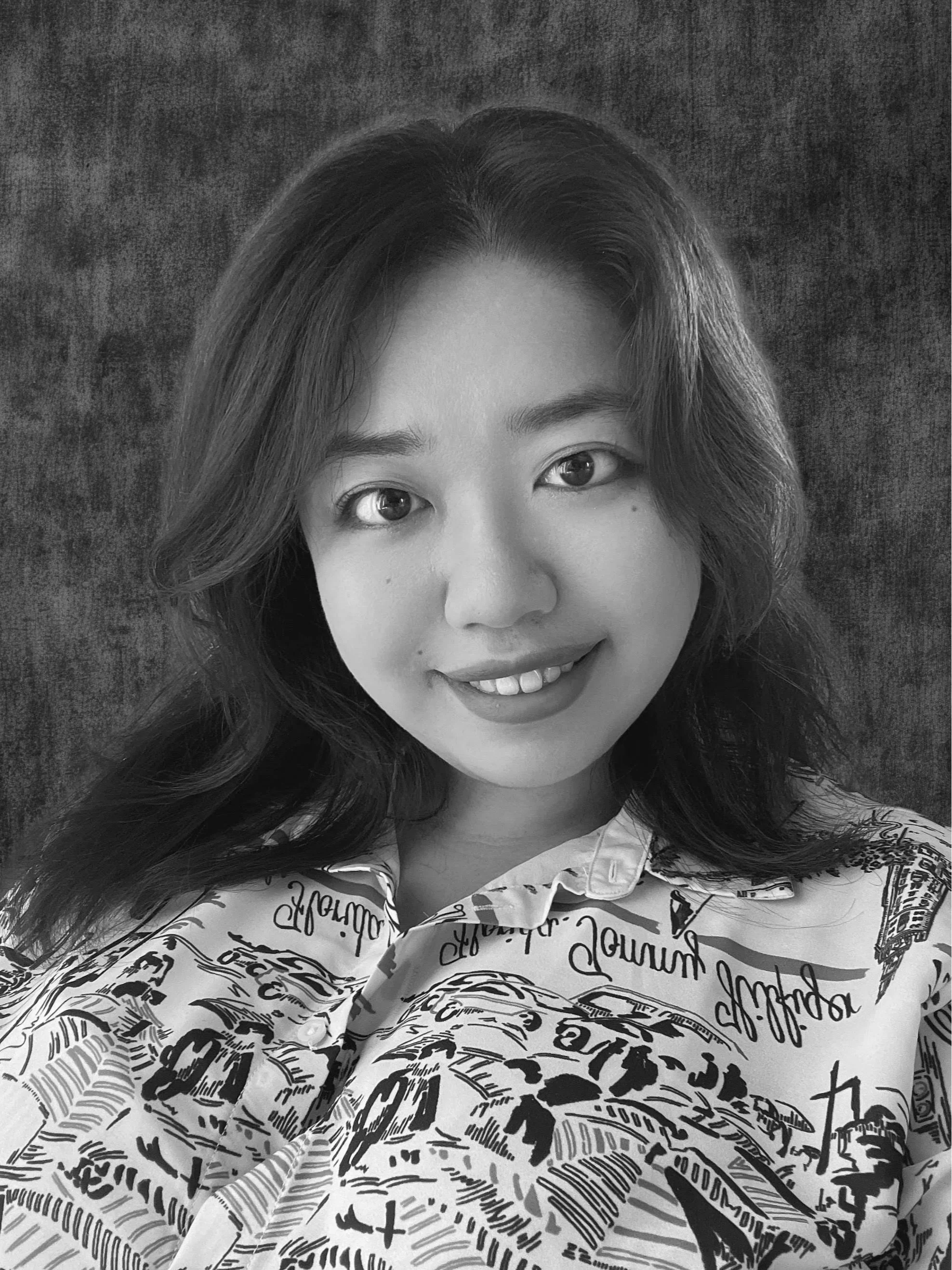 Portrait of a young woman with shoulder-length wavy brown hair, smiling, wearing a white shirt with black and green graphic print, against a dark textured background.