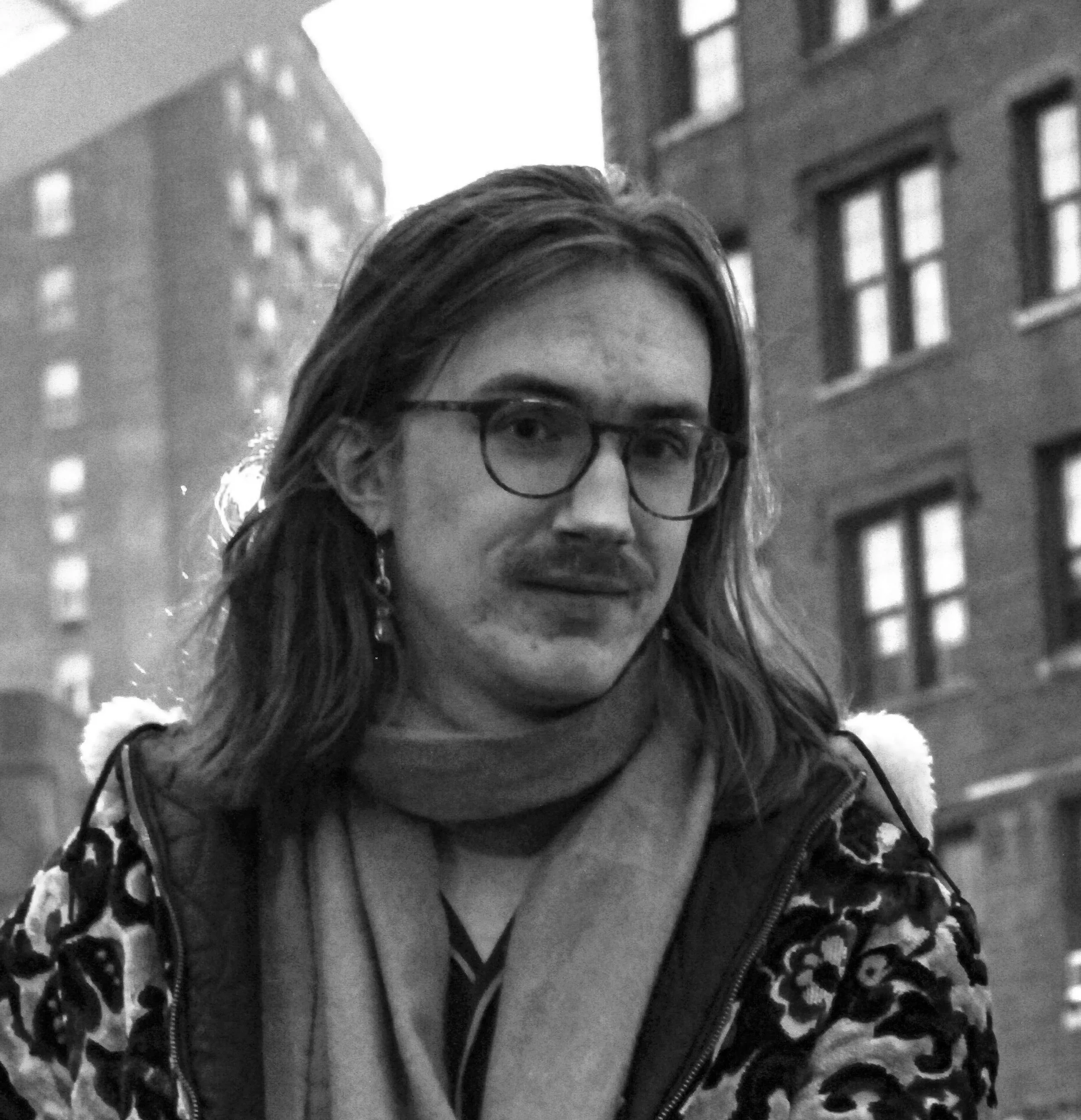 Black and white portrait of a smiling young man with glasses and a beard, wearing a floral short-sleeve shirt, standing outdoors near water with trees in the background.