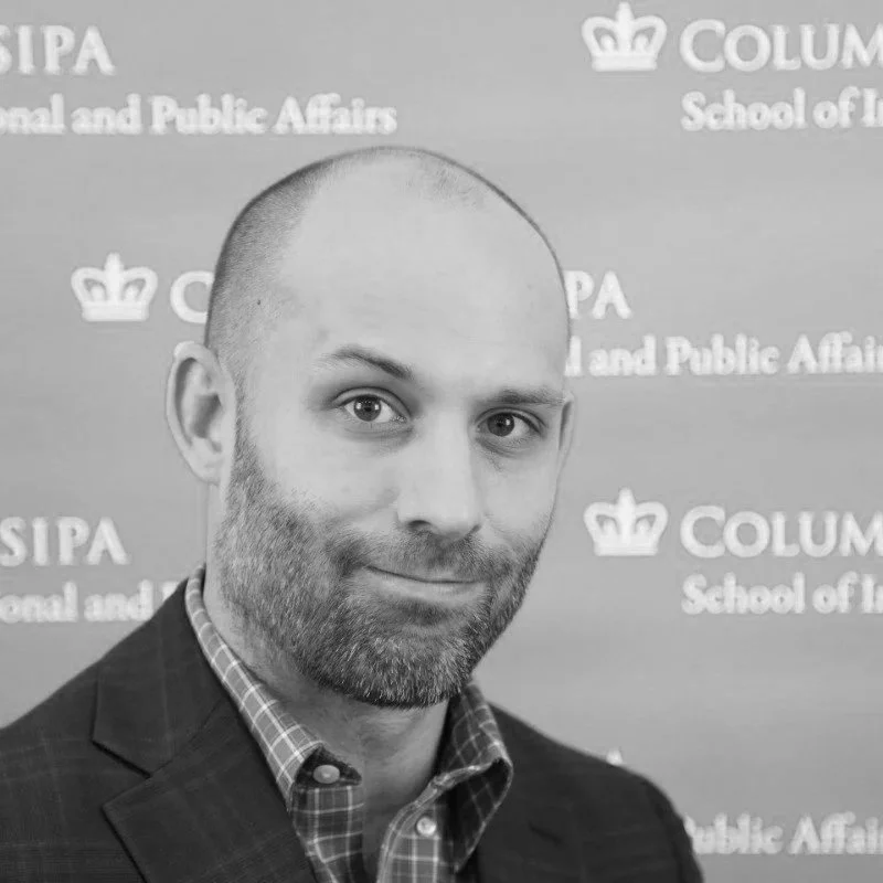 Portrait of a man with a beard and short hair, wearing a dark jacket over a blue checkered shirt, standing in front of a blue backdrop with white text and logos for Columbia School of International and Public Affairs.