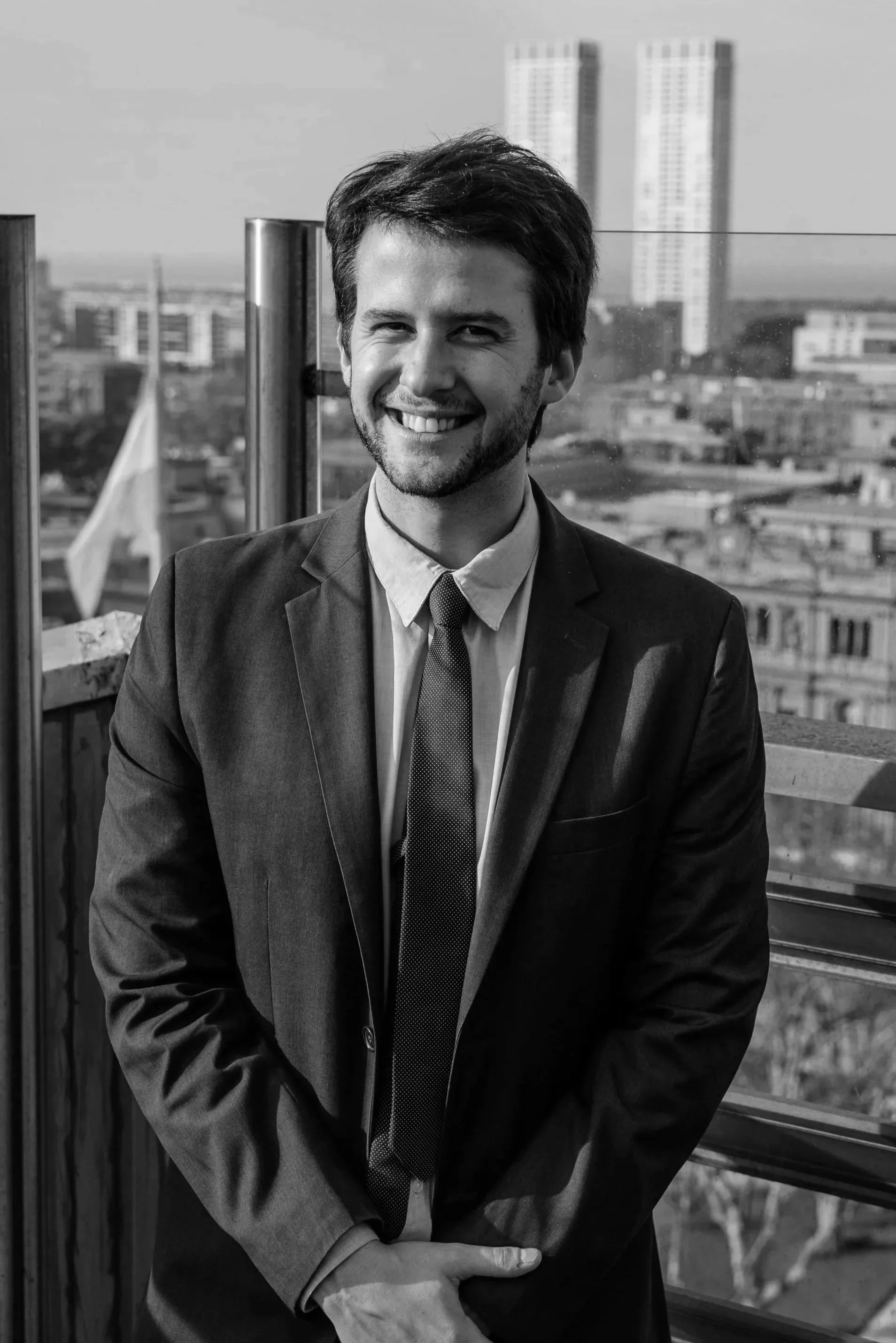 A man smiling in a business suit standing on a rooftop with a cityscape in the background.