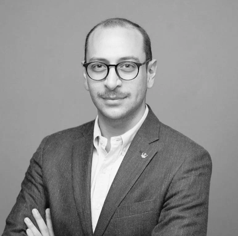 A man wearing glasses, a navy blue blazer, and a light pink collared shirt, posing with arms crossed against a plain gray background.