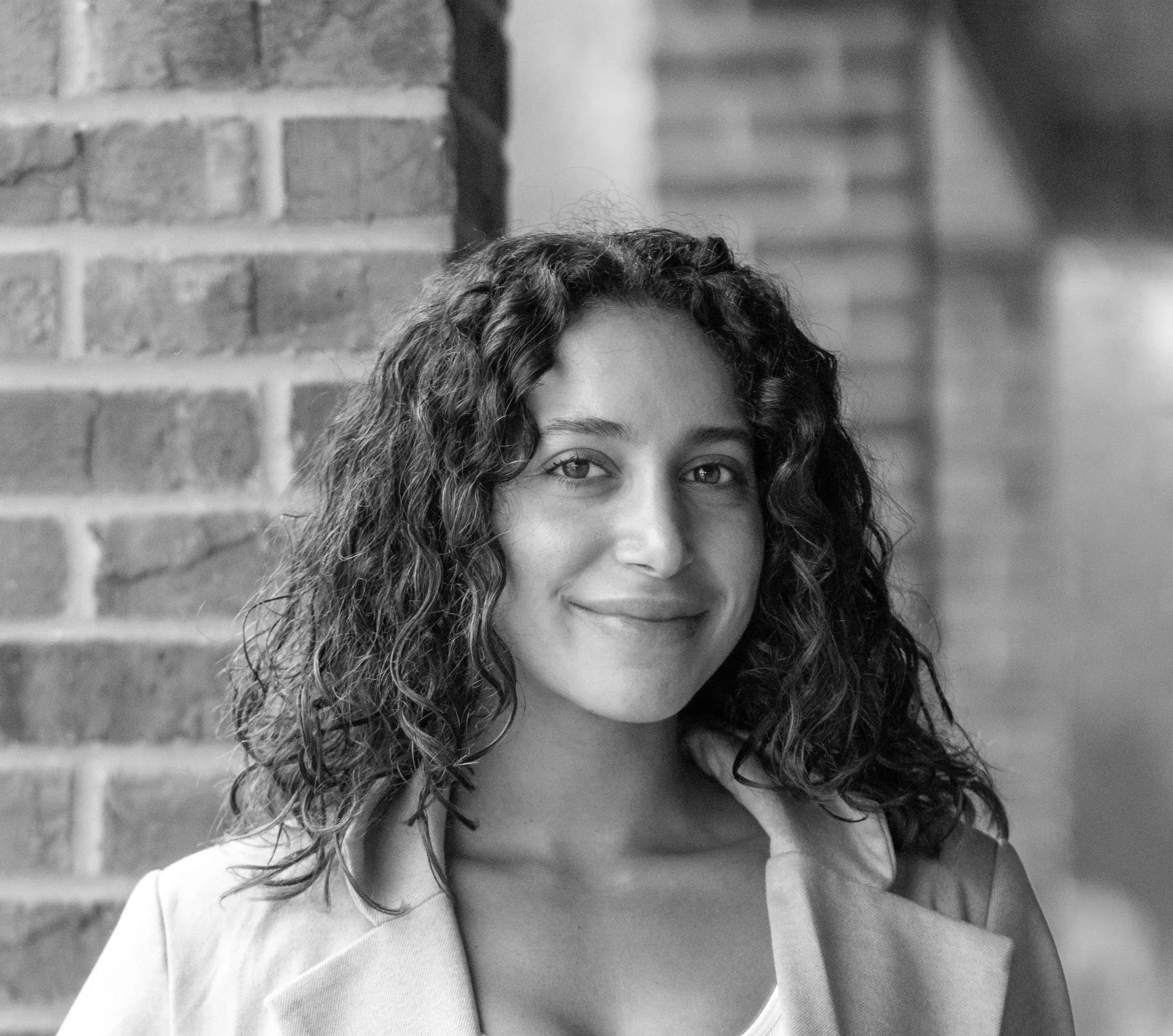 A woman with curly hair smiling, standing near a brick wall with stairs in the background.