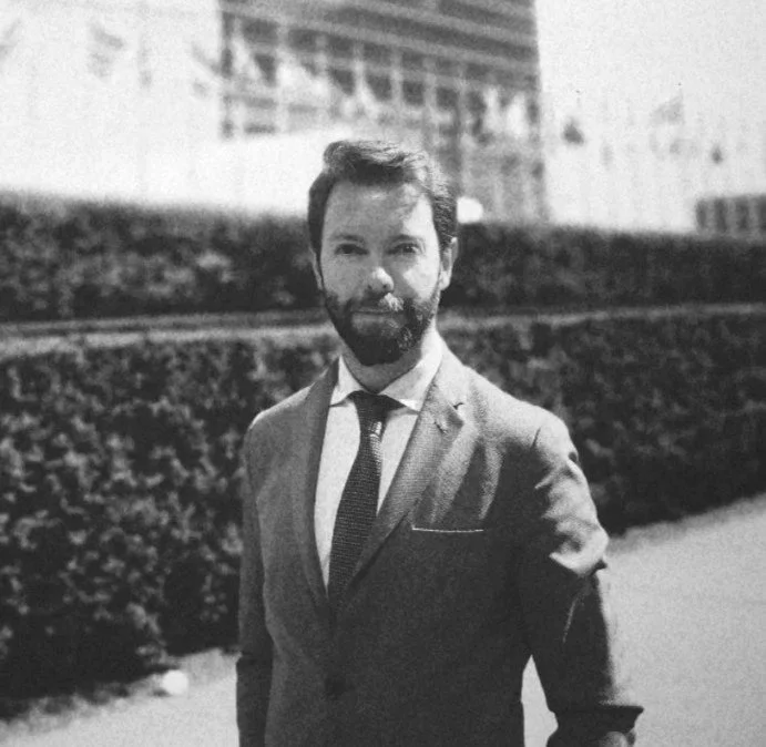 A black and white photo of a man in a suit standing outdoors with buildings and a hedge in the background.