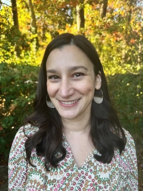 A woman with dark hair smiling outdoors, wearing a patterned blouse and hoop earrings, with trees and autumn foliage in the background.