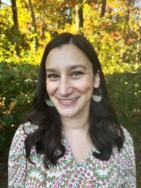A woman with long dark hair and earrings smiling outdoors with colorful trees and leaves in the background.