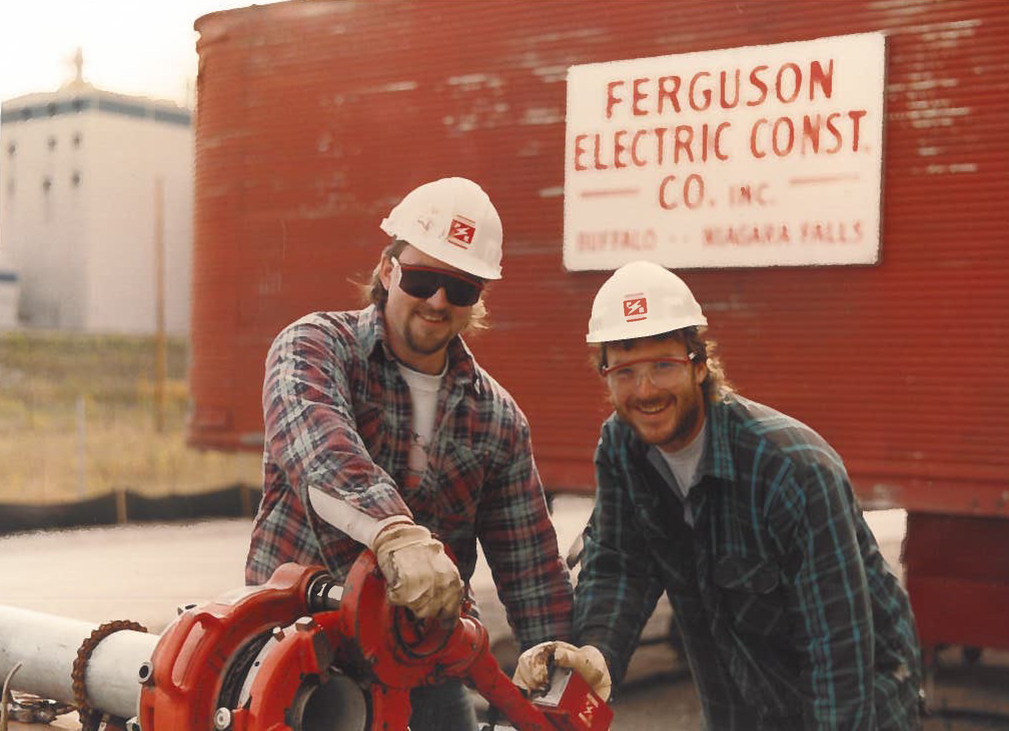 Two men wearing safety helmets and glasses working with a pipe cutting machine outdoors, with a red building and a white sign that says Ferguson Electric Const Co.