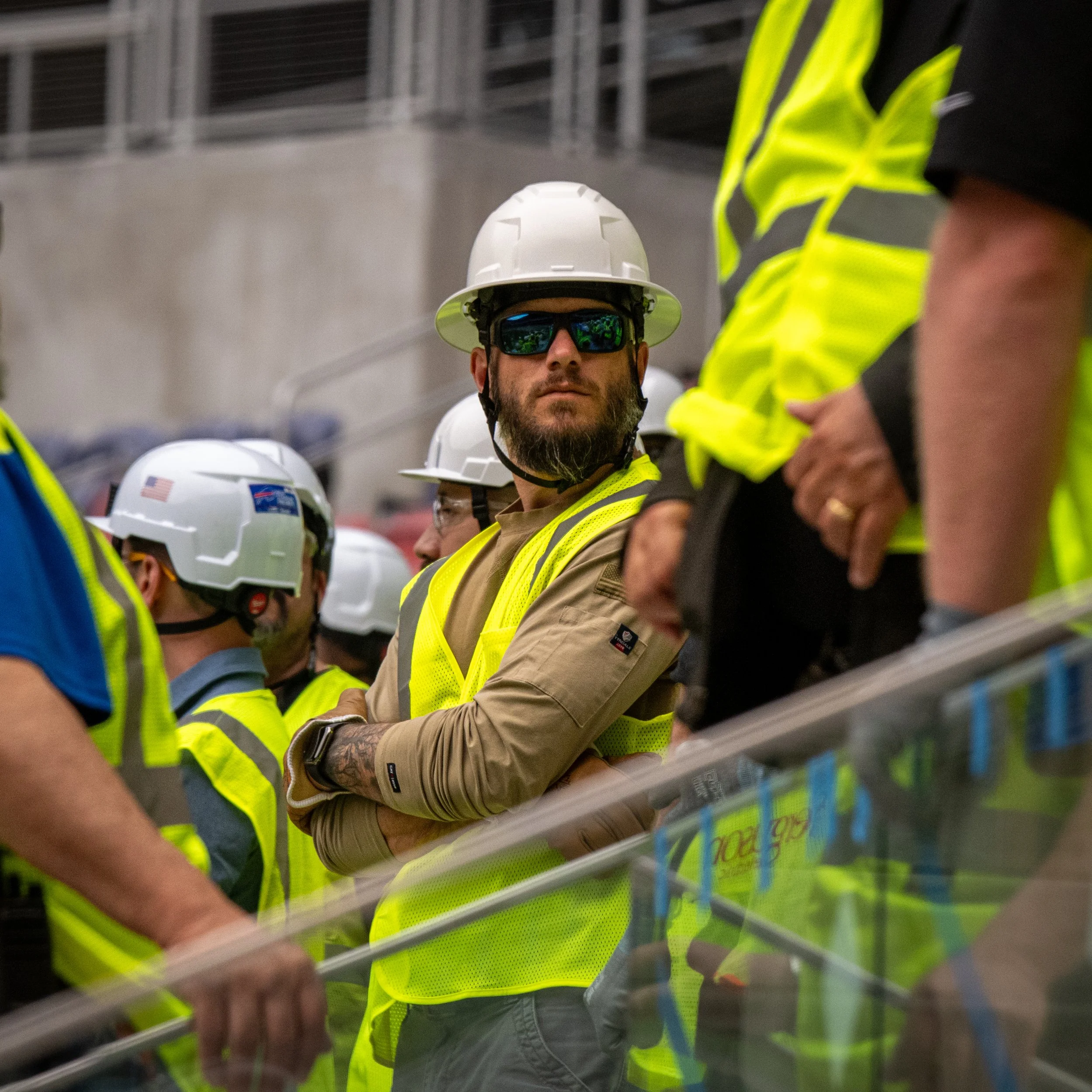 A person wearing a white hard hat, reflective sunglasses, and a yellow safety vest, shaking hands with another individual who is partly visible and also in safety gear, inside an industrial or construction setting.