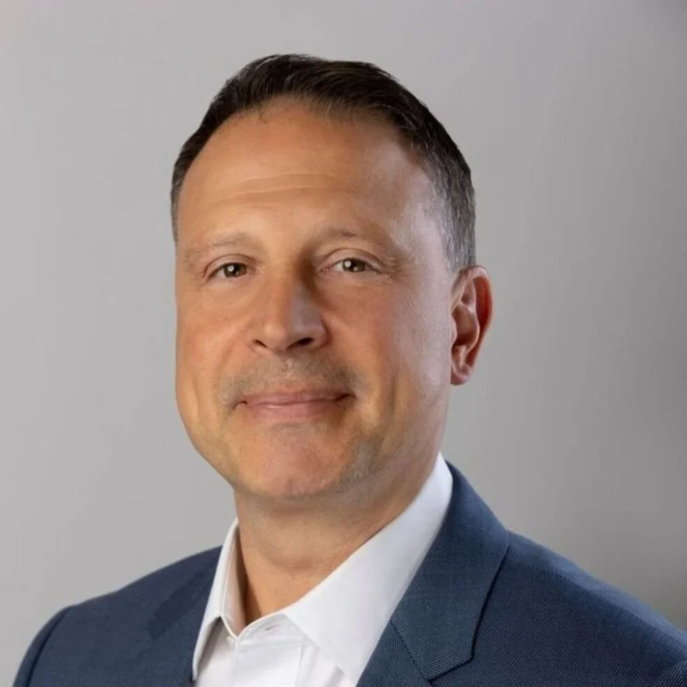 Professional headshot of a man in a dark blue suit and white shirt against a gray background.