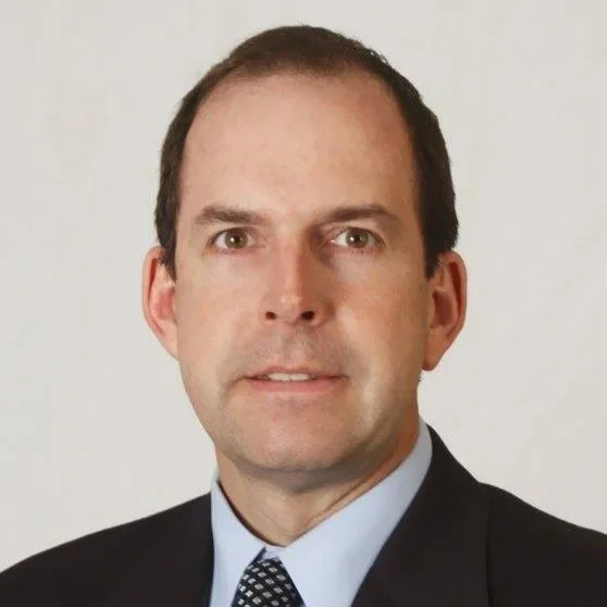 Headshot of a man with short dark hair, wearing a dark suit, light blue shirt, and patterned tie, against a plain light background.