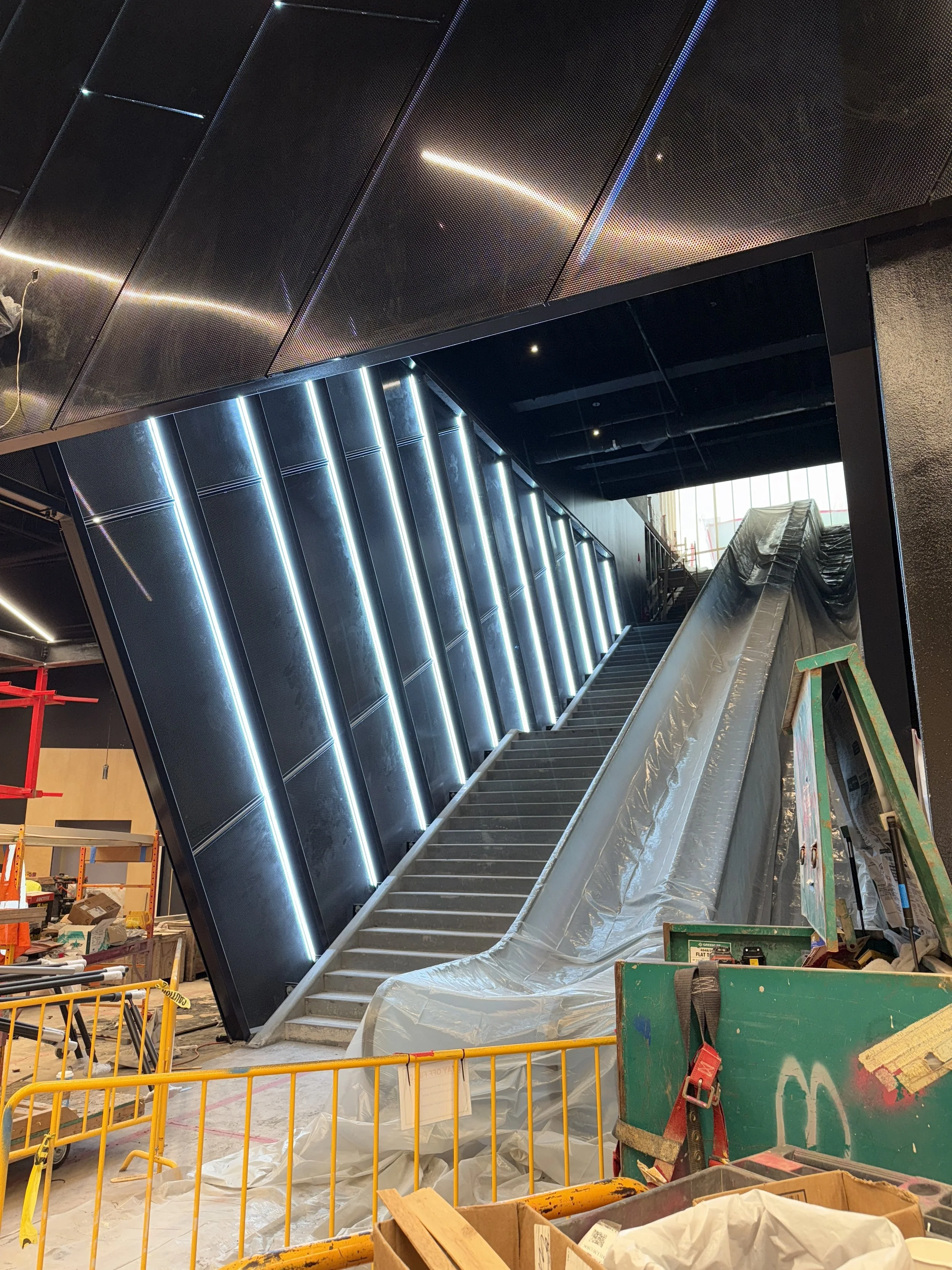 Interior of a building under construction with a staircase and modern lighting. Construction tools and materials are scattered around, and safety barriers are in place.