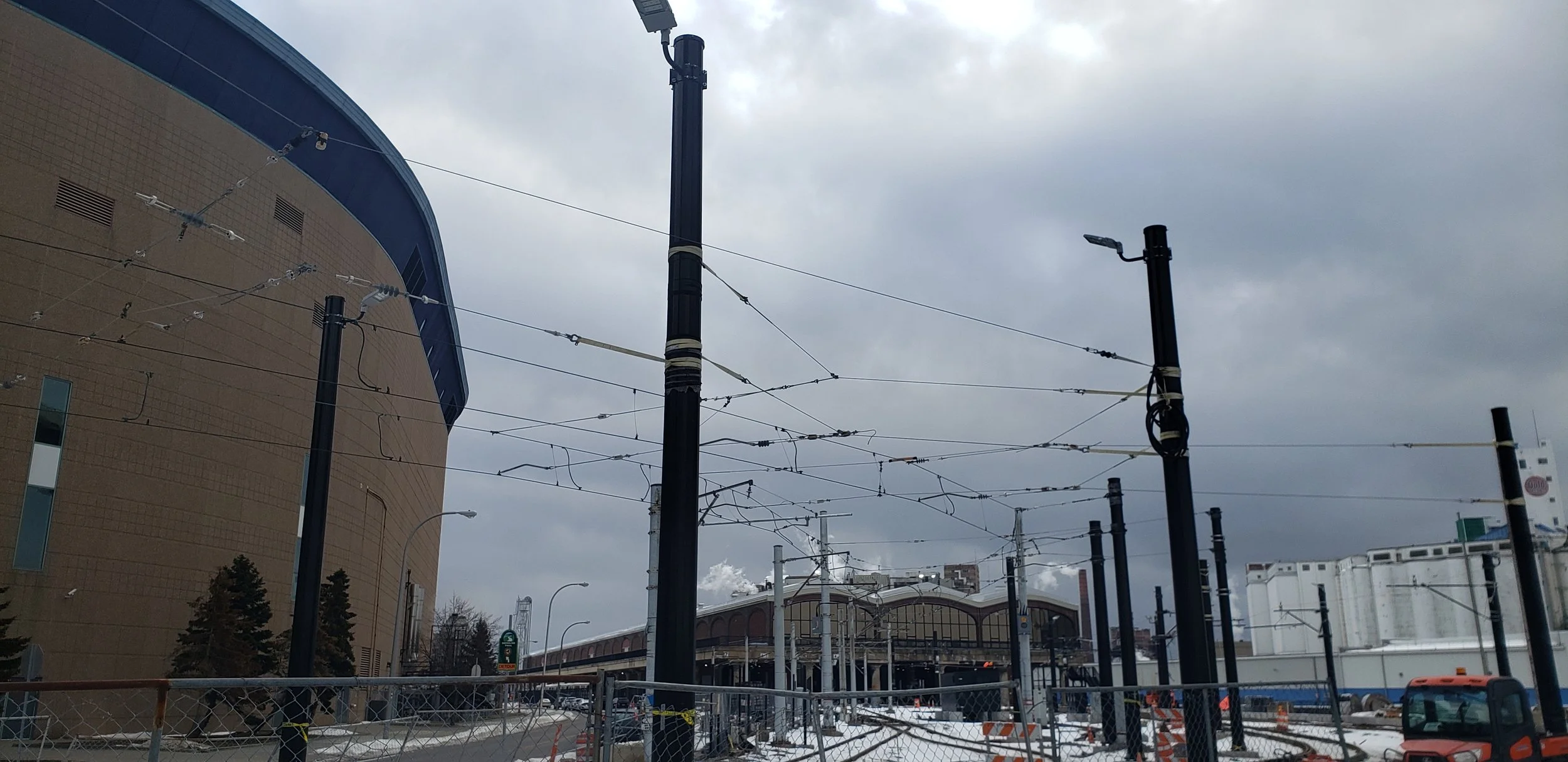 Construction site with utility poles and overhead wires, surrounded by a fence, near a modern building under cloudy sky.
