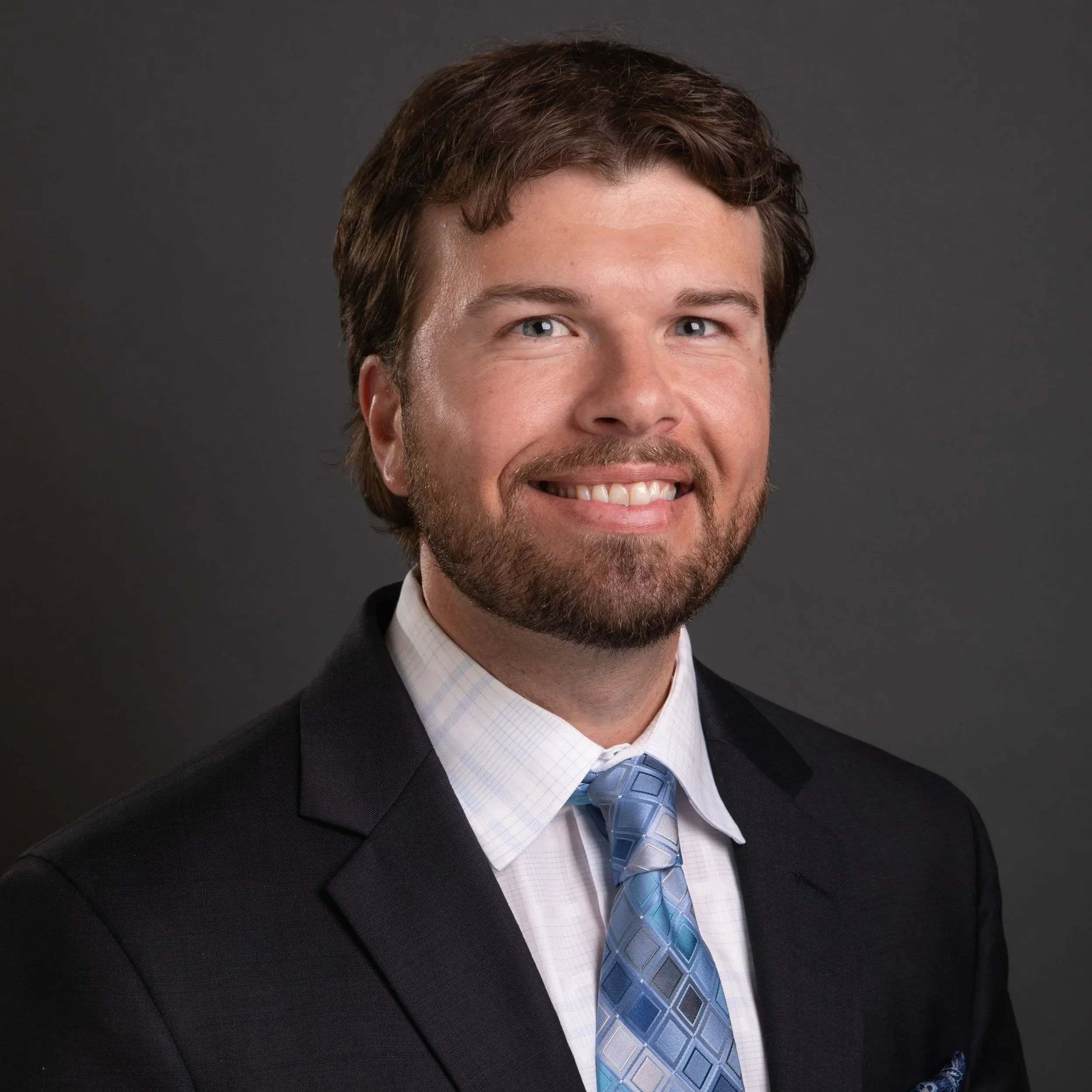 Close-up of a man smiling, wearing a dark suit, light blue checkered shirt, and a blue patterned tie, against a gray background.