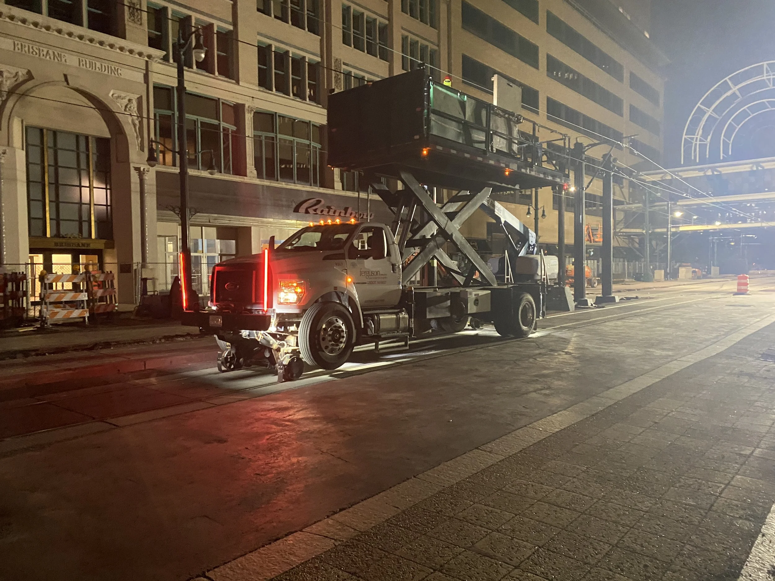 Night scene of a city street with a construction lift vehicle parked on the side. The lift is raised with a person in a yellow helmet visible. There are buildings with large windows and decorative facades along the street. Streetlights illuminate the