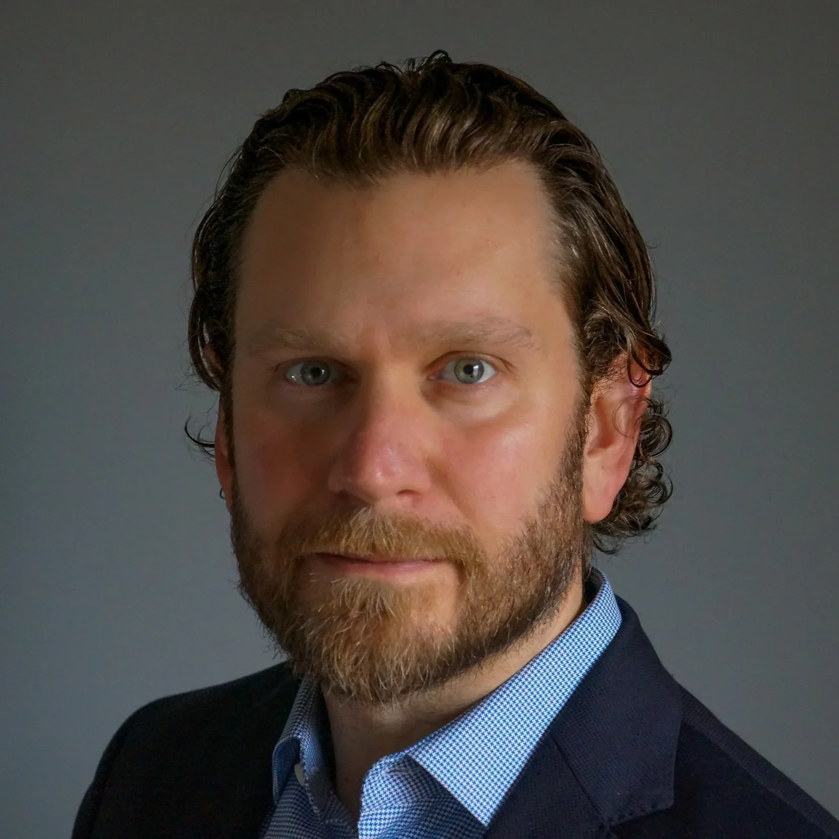 Close-up of a man with light skin, green eyes, brown curly hair, and a beard, wearing a dark jacket over a blue checkered shirt, against a gray background.