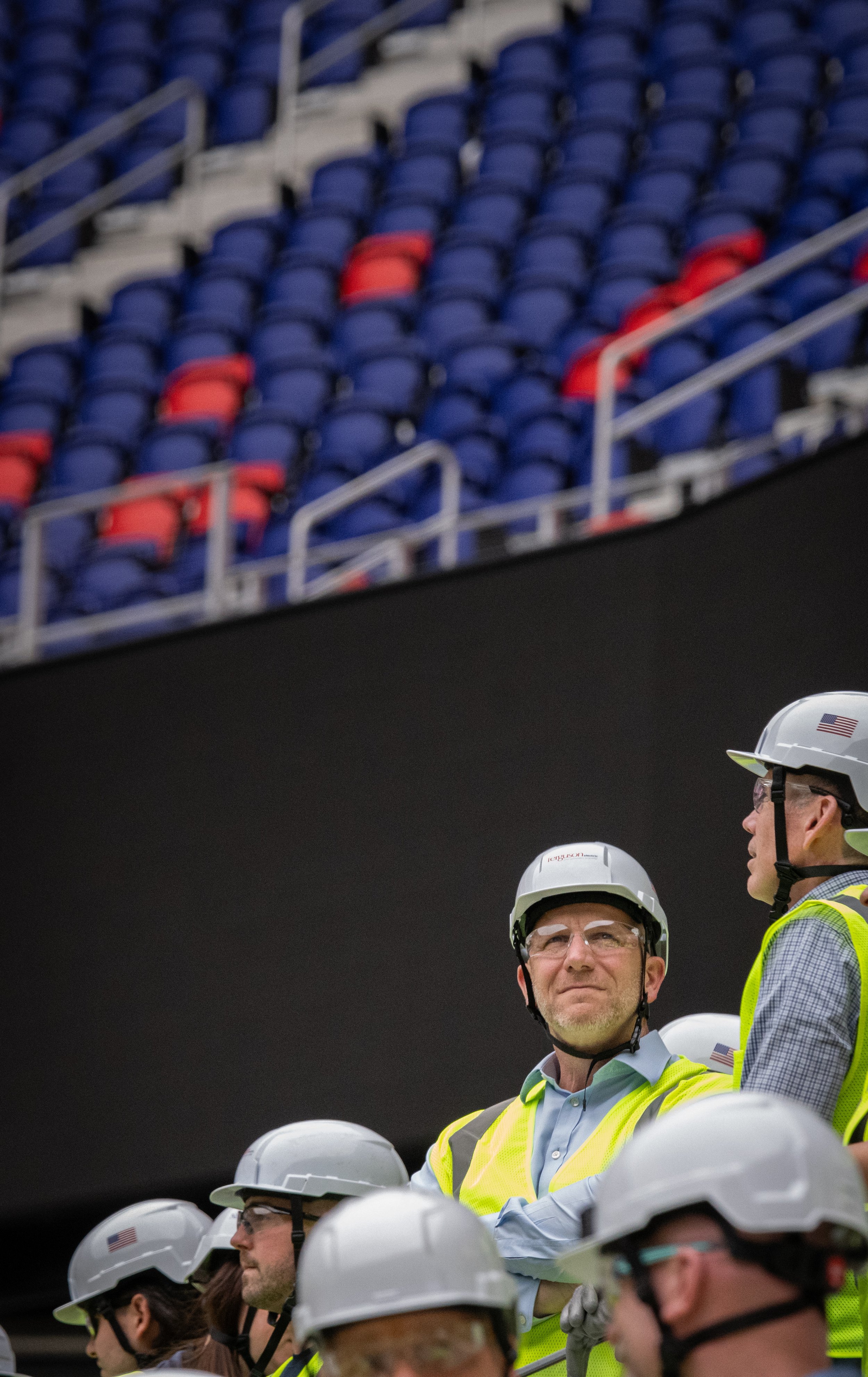 Group of people wearing safety helmets and yellow vests, standing in formation at a construction site. Some are looking ahead, with a cloudy or indoor background. The image captures a moment of safety and teamwork.