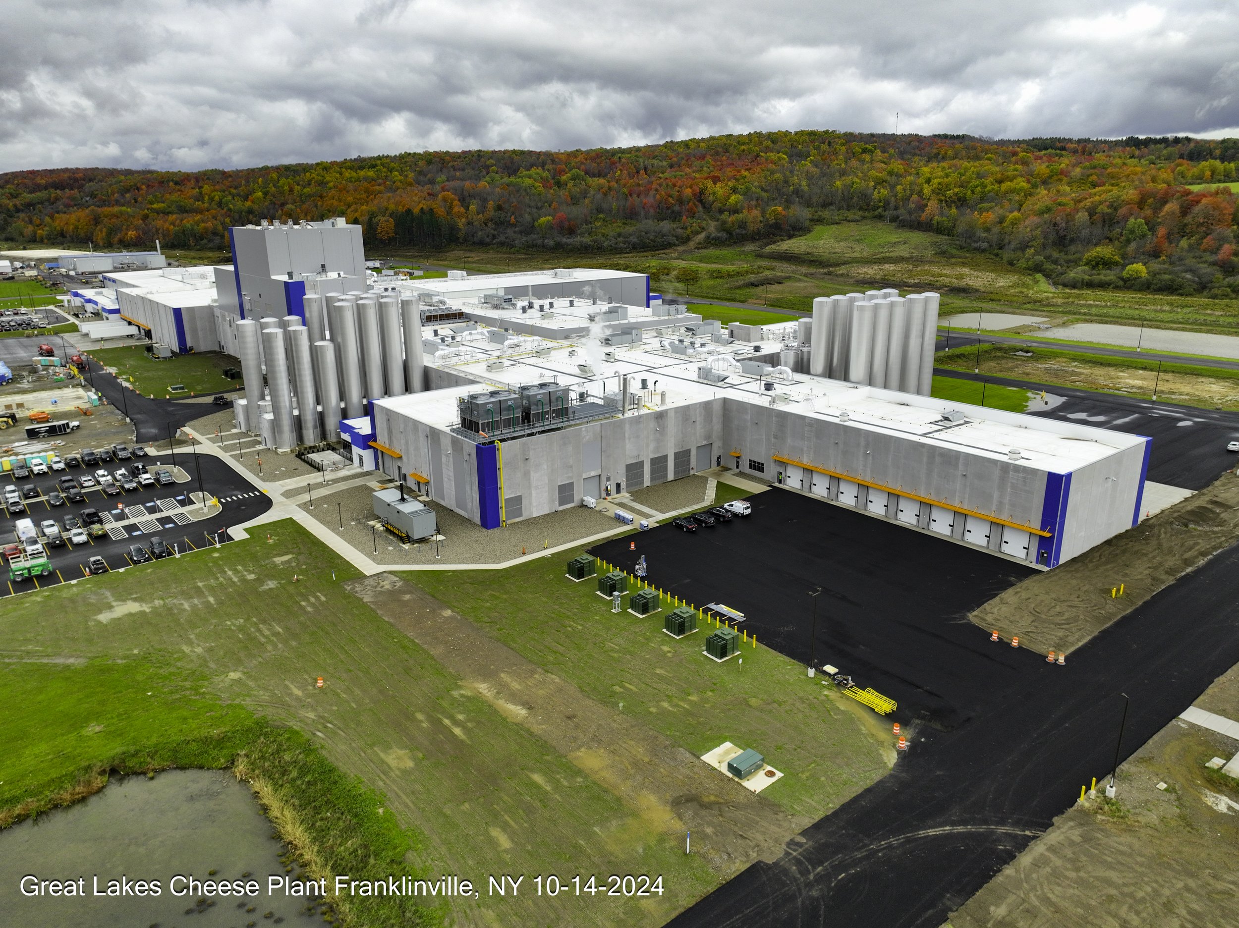 Aerial view of a large cheese processing plant with white buildings, silver silos, parking lot, and lush green surroundings with a hill covered in colorful autumn trees in the background.
