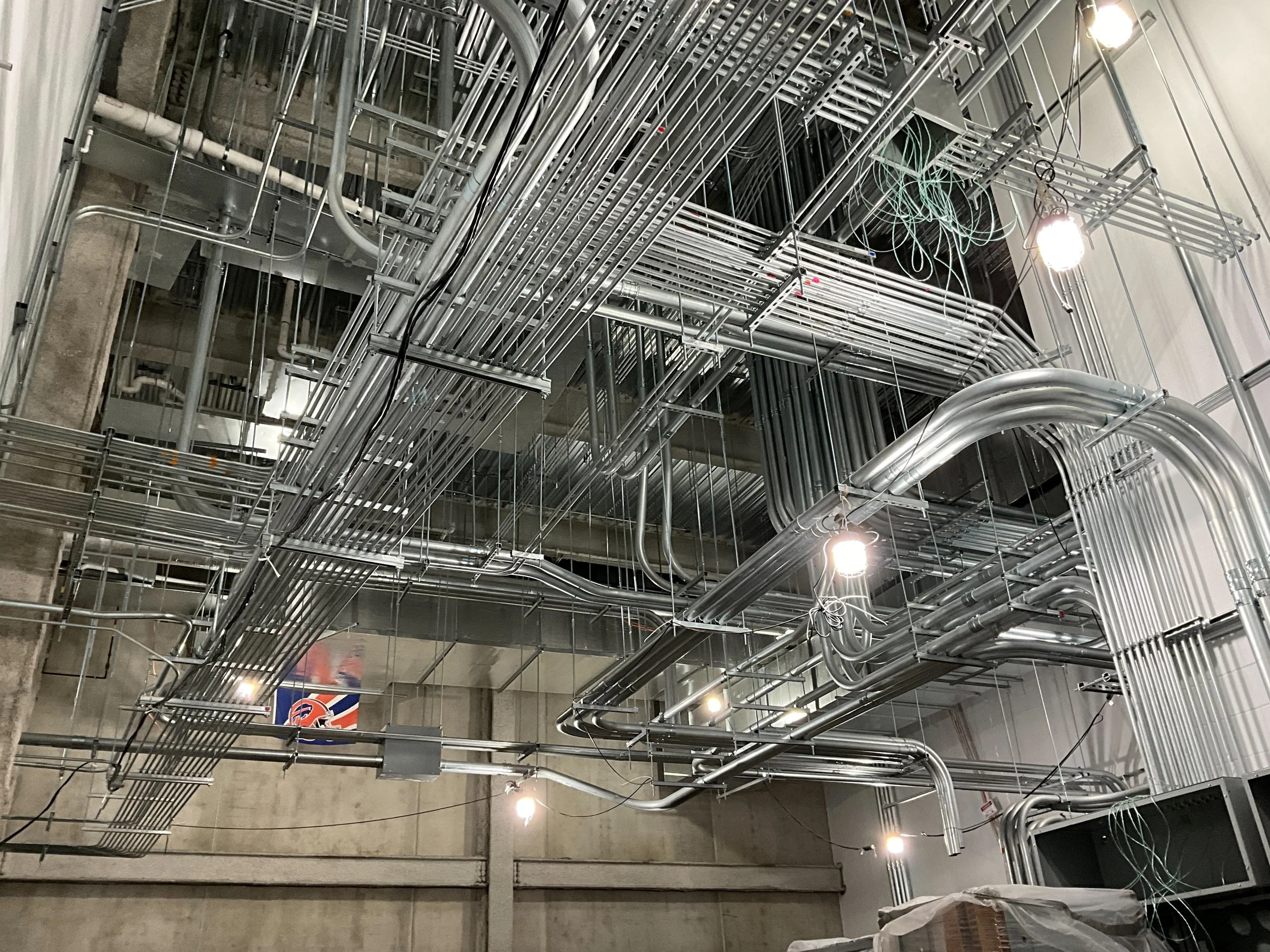 An industrial space with a complex network of metal pipes and cables on the ceiling, illuminated by bright lights.