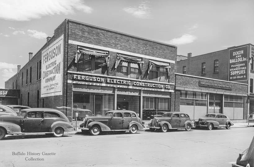 Black and white photo of a two-story brick building labeled Ferguson Electric Construction Co. with cars parked in front and awnings over the windows. Sign on the side of the building mentions Ferguson Electric Company.