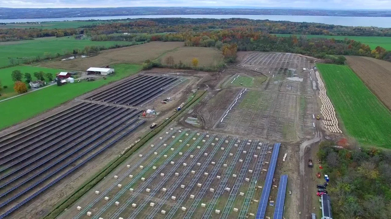 Aerial view of a solar farm under construction on farmland, with rows of solar panels being installed on the fields, adjacent to green fields, trees, and a body of water in the background.