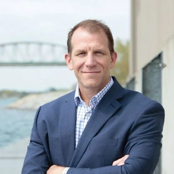 Man in a blue blazer and checked shirt standing outdoors near a body of water with a bridge in the background.
