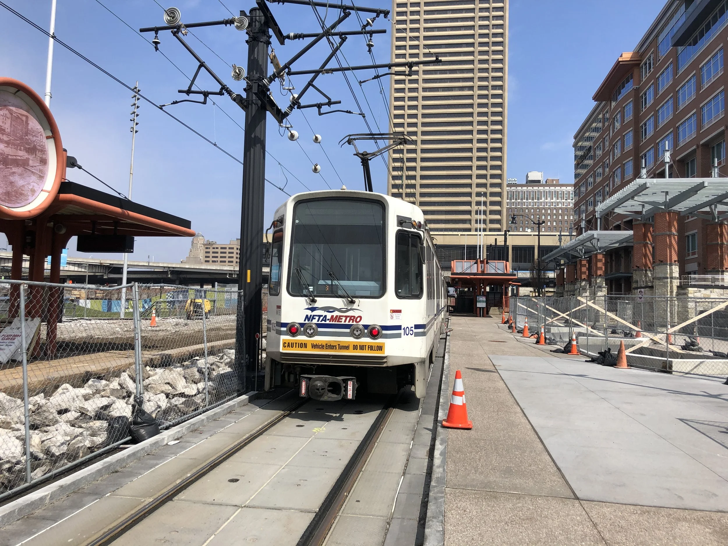 A construction site in an urban area with a small light rail vehicle in the foreground. The vehicle has a sign that reads "CAUTION Vehicle Enters Tunnel DO NOT FOLLOW." Orange cones line the sidewalk with a fenced-off area containing rocks and constr