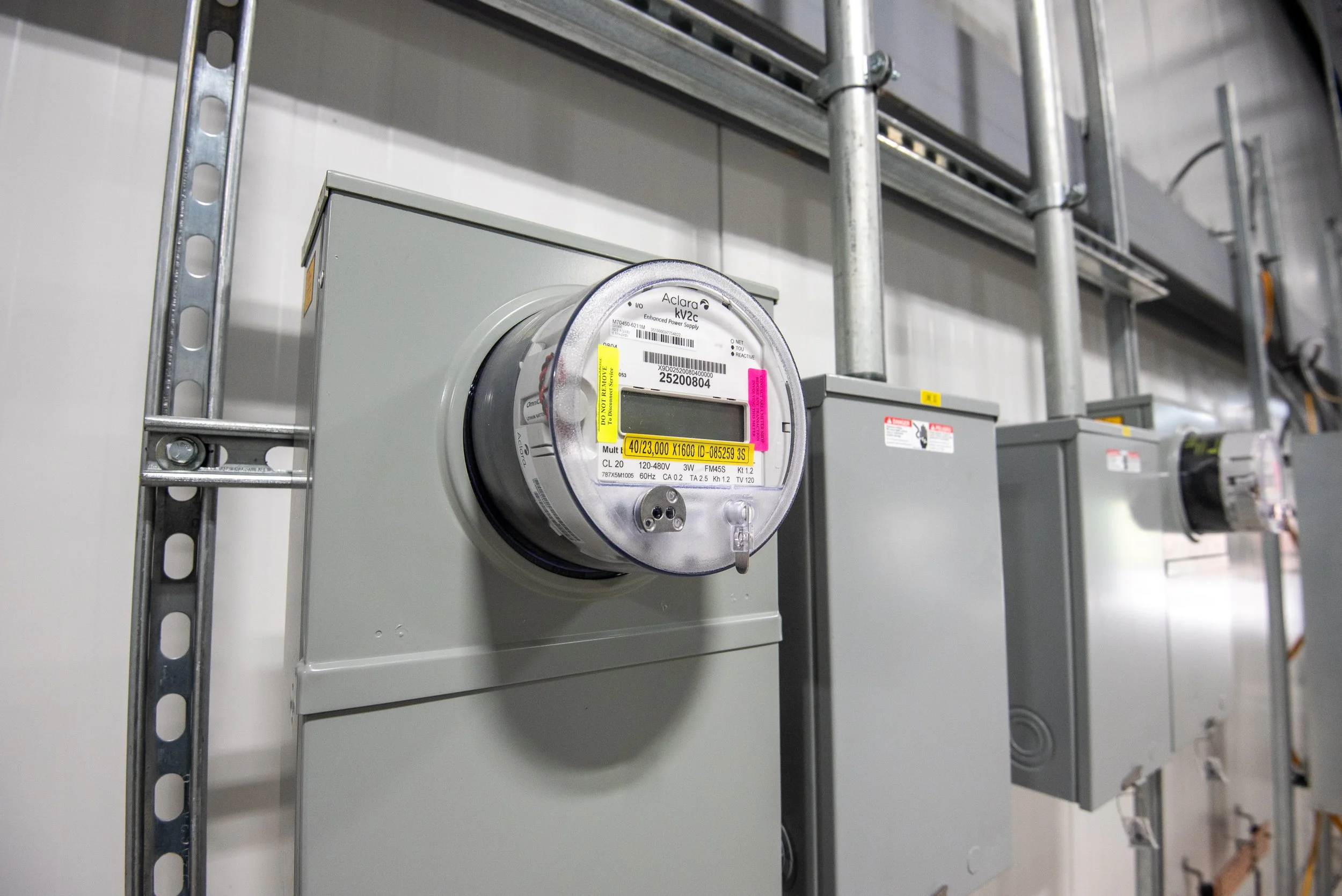 Electrical meter mounted on a gray electrical box attached to a metal shelving unit in an industrial setting.