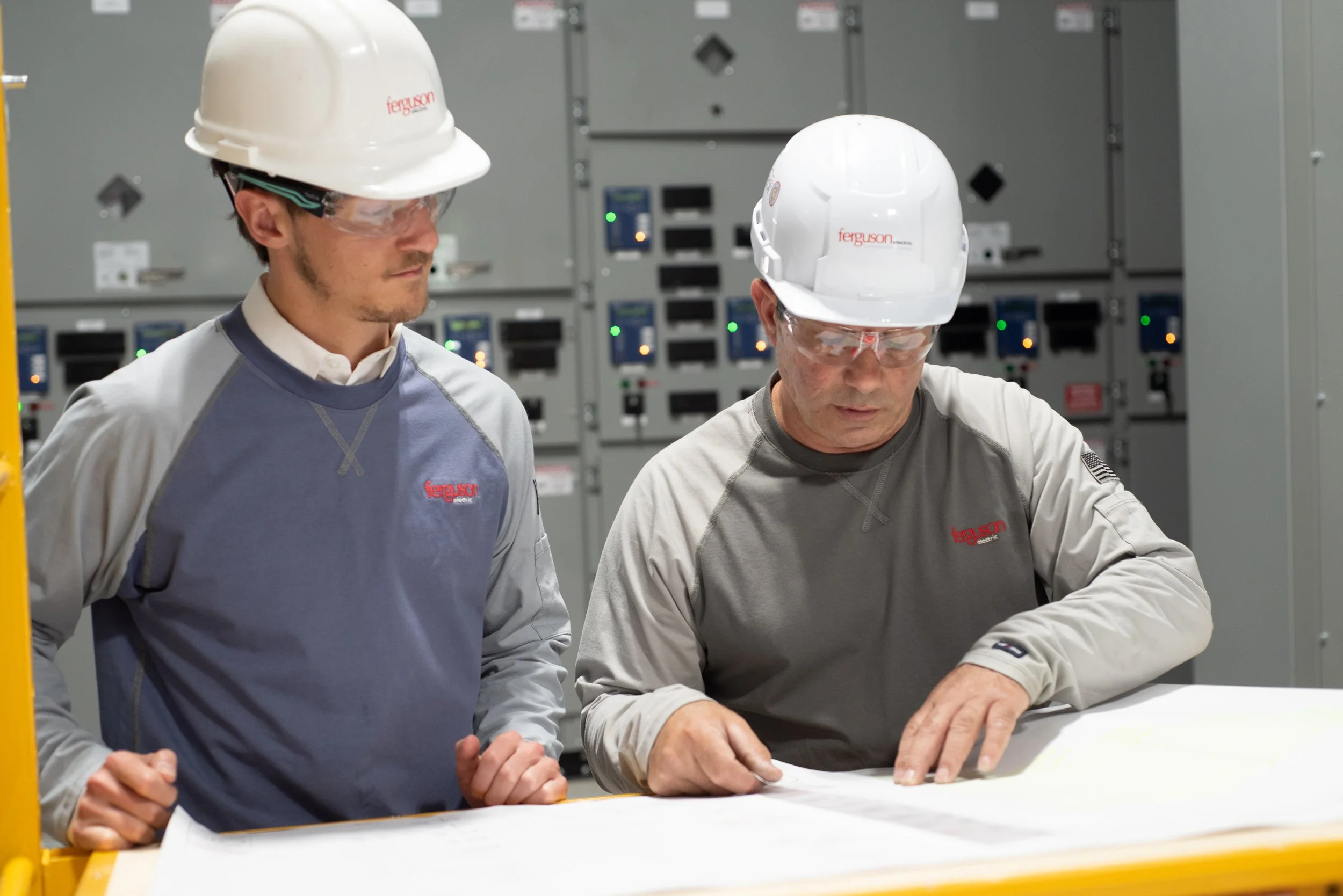 Two men in safety helmets and protective glasses working together at a table with blueprints or plans in a control room with electrical panels in the background.
