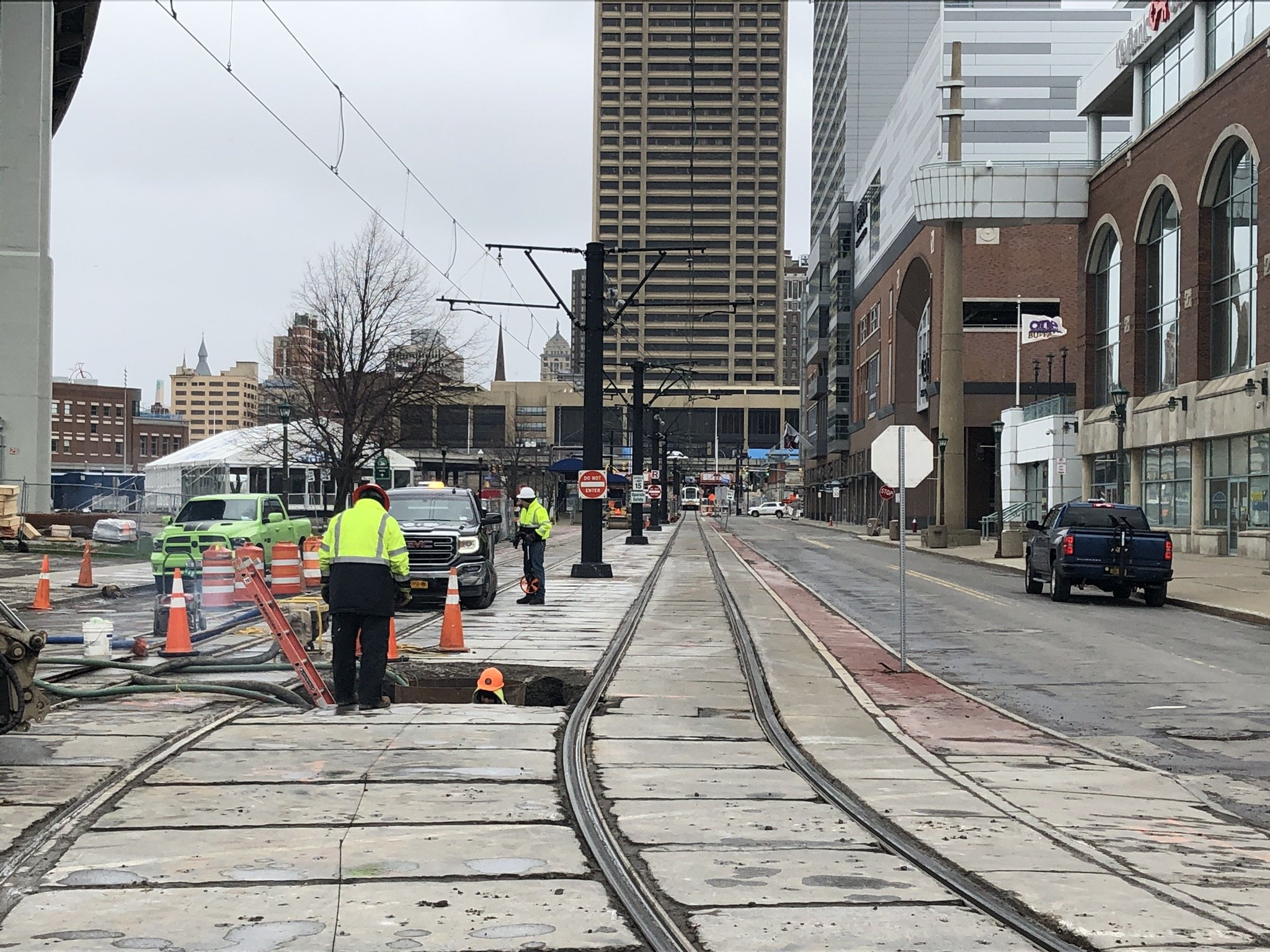 City street with tracks for a tram or light rail, construction workers repairing or installing the tracks, surrounded by tall buildings and some trees, overcast sky.