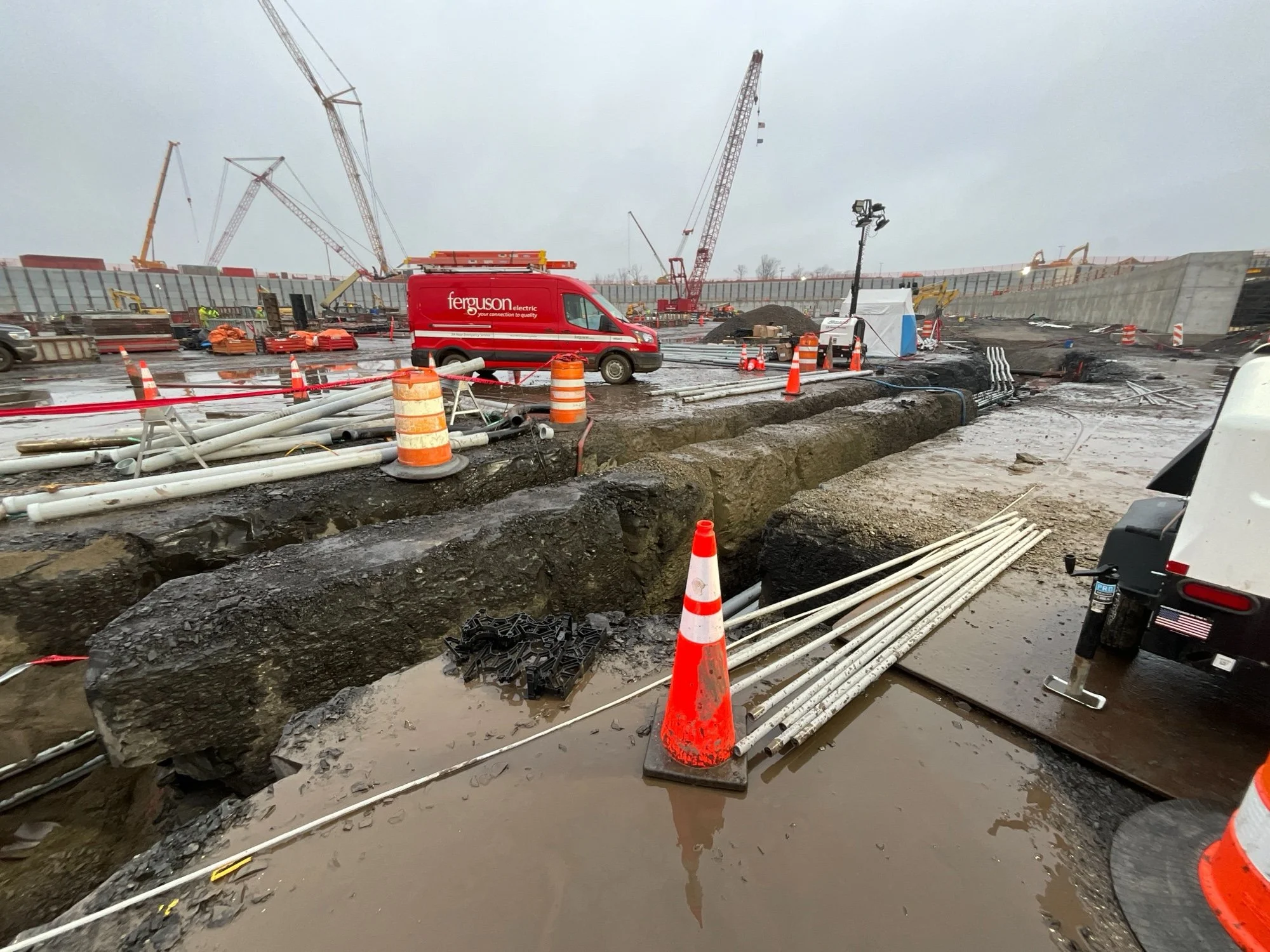 Construction site with large trenches, pipes, and construction equipment, including cranes and a red van, on a cloudy day.