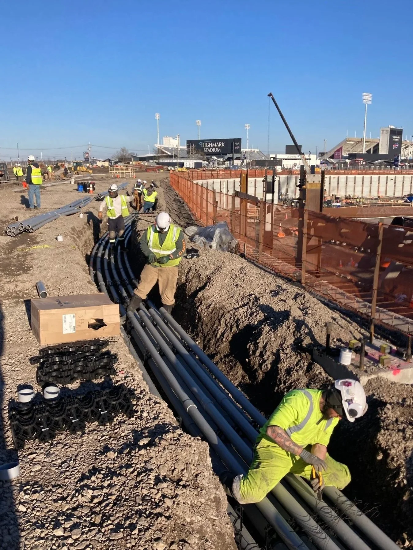 Construction workers installing underground pipes at a stadium construction site on a clear day.